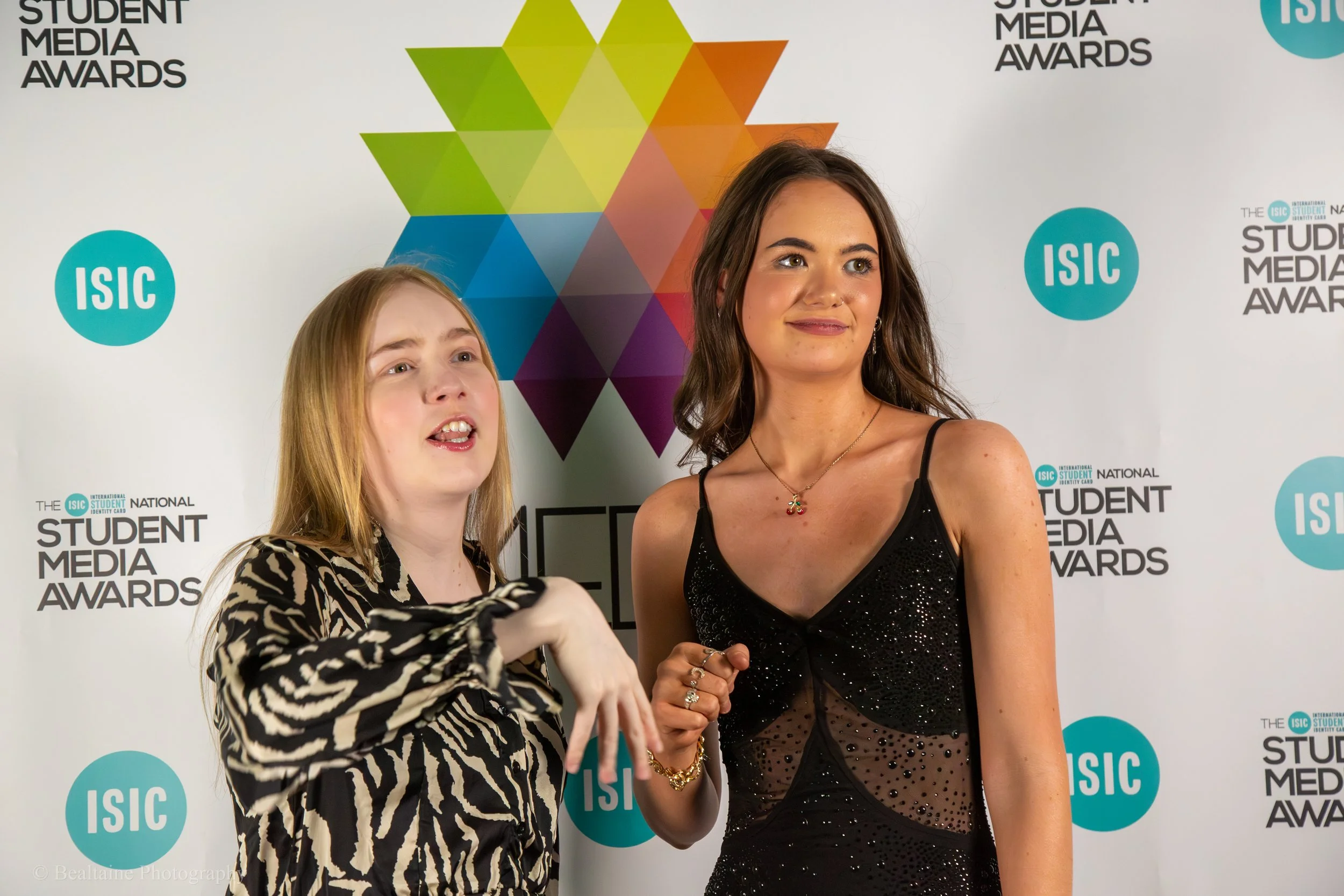 Two women at a media awards event, standing in front of a backdrop with colorful geometric shapes and the logos 'ISC' and 'Student Media Awards'.