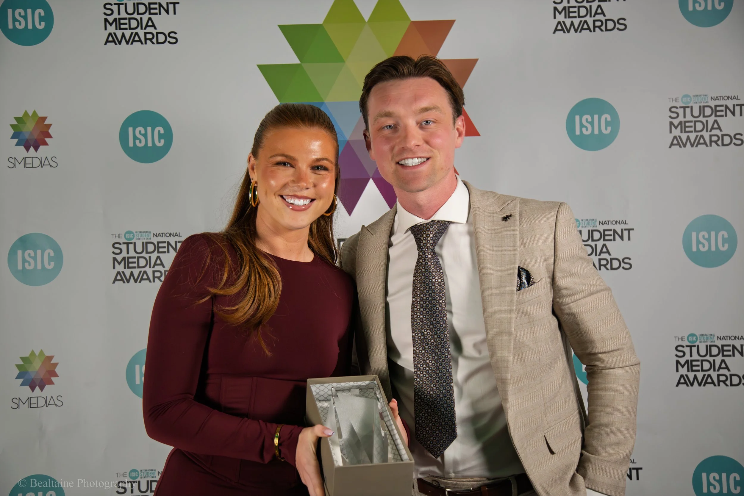 A woman and a man smiling at the camera at the Student Media Awards event, with a backdrop displaying logos for ISIC and student media awards.