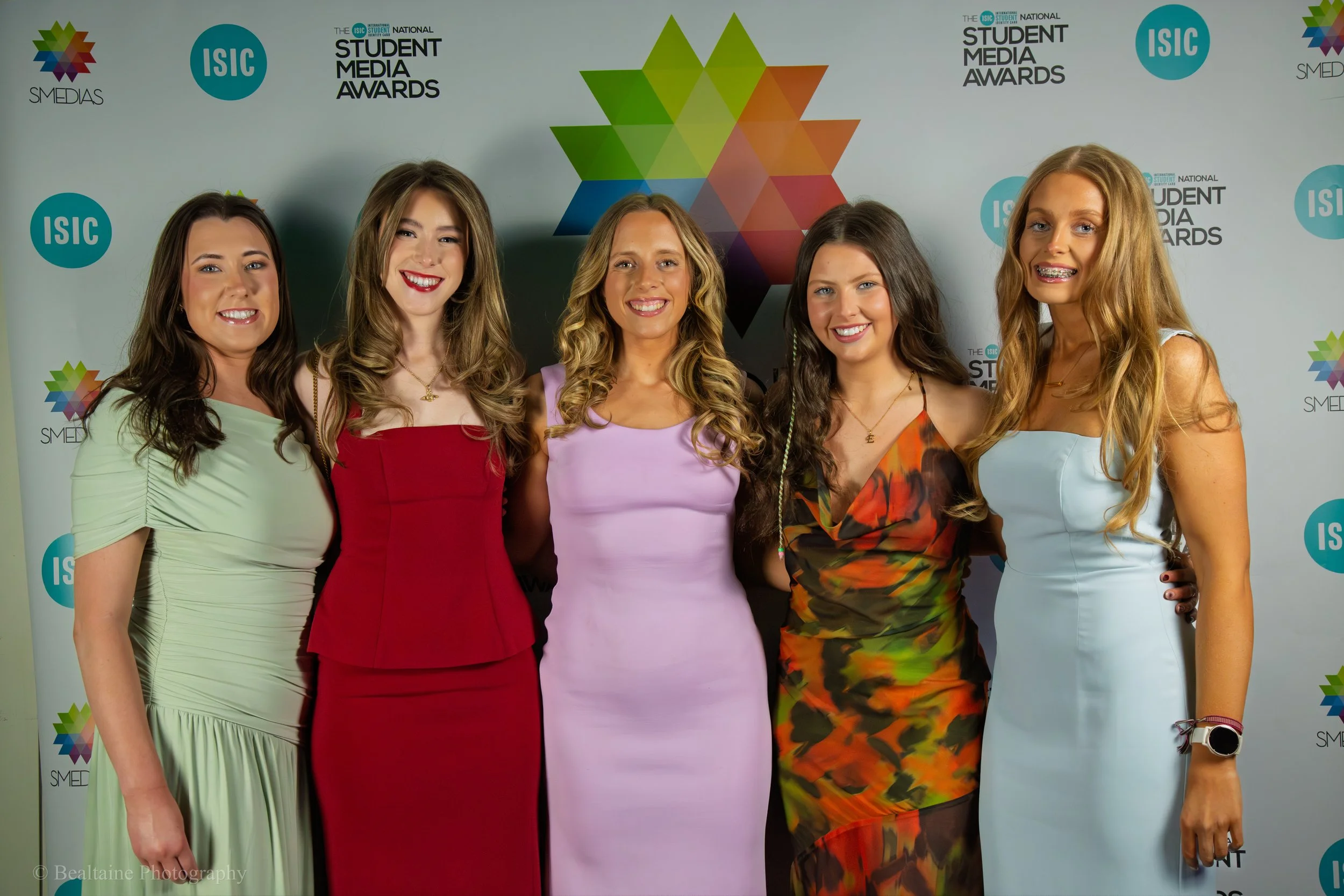 Five young women in colorful dresses smiling at a media awards event, standing in front of a backdrop with logos and geometric shapes.