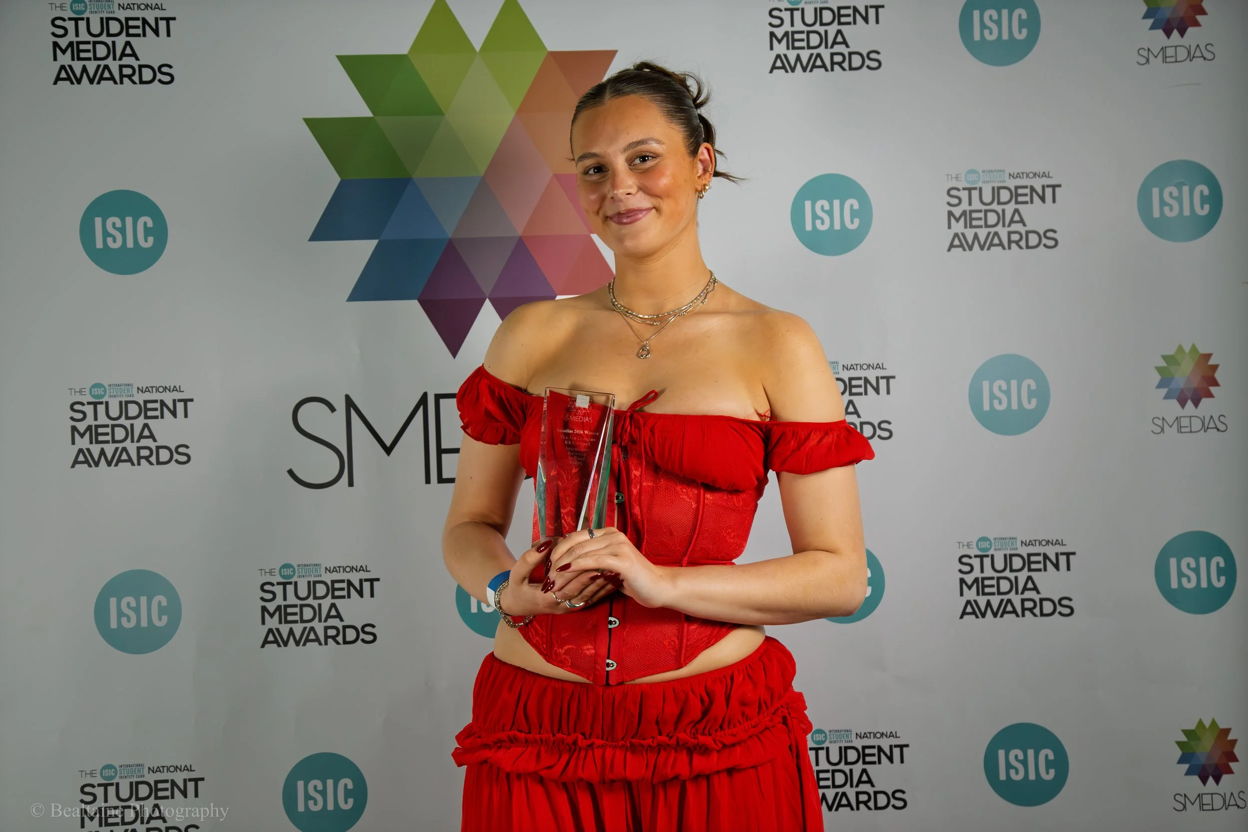 A woman in a red off-the-shoulder dress holding a clear award trophy at the Student Media Awards event, standing in front of a backdrop with logos and text related to the awards.