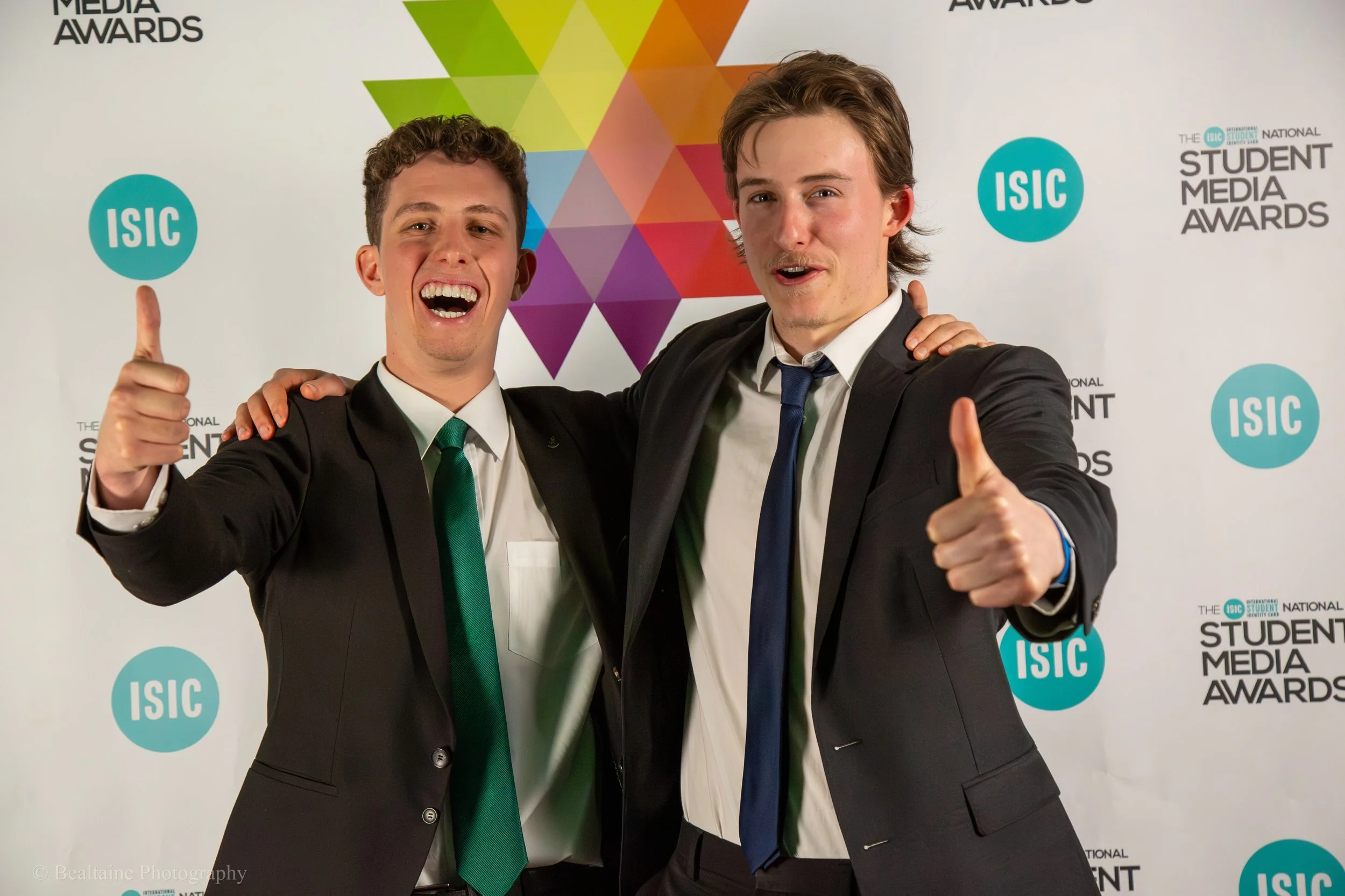 Two young men in suits smiling and celebrating at the Student Media Awards, with one giving a thumbs-up. Behind them is a white backdrop with colorful geometric shapes and the logos for ISIC and the awards.
