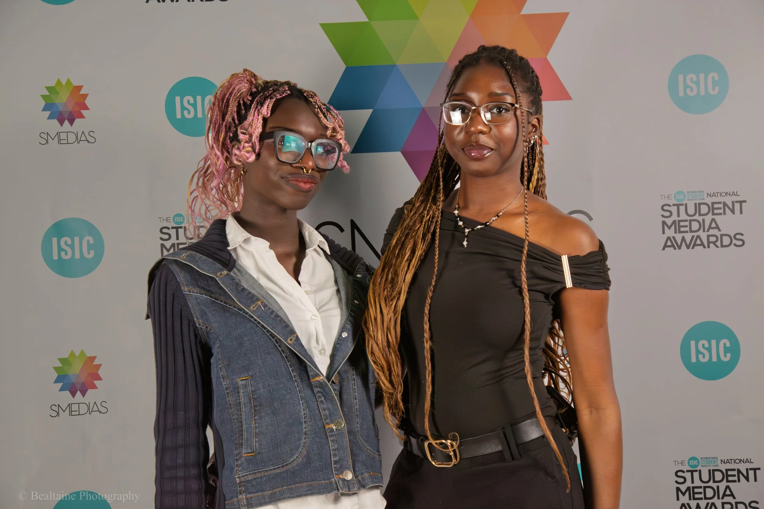 Two women standing in front of a backdrop with multicolored geometric logo and text that says "STUDENT MEDIA AWARDS." The woman on the left has short, pink, and purple braided hair, glasses, and is wearing a denim jacket over a white shirt. The woman