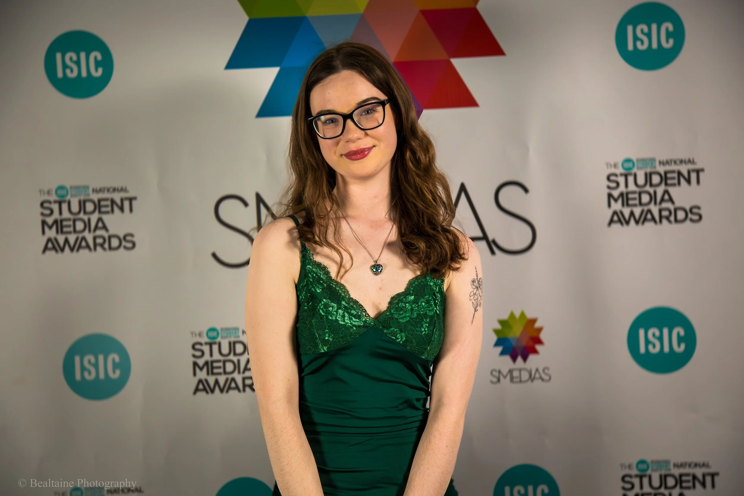 A young woman with long brown hair, glasses, and red lipstick, wearing a green lace dress with spaghetti straps, stands in front of a backdrop with logos for ISC and the Student Media Awards.