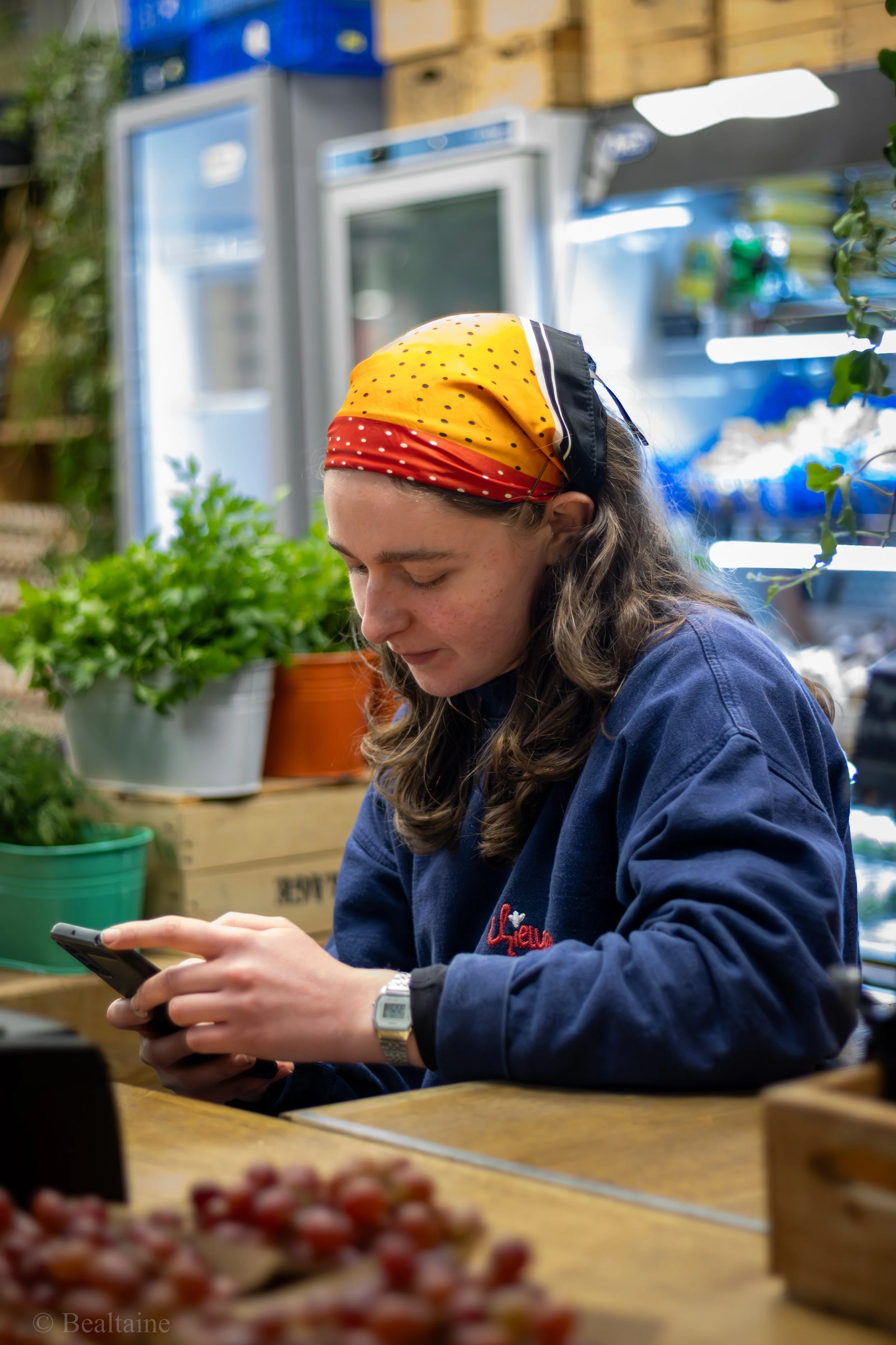 A woman with long, wavy hair wearing a yellow, red, and black patterned headscarf and a navy blue jacket, sitting at a wooden table in a greenhouse or garden center, looking at her smartphone. Behind her are various potted plants, and in the backgrou