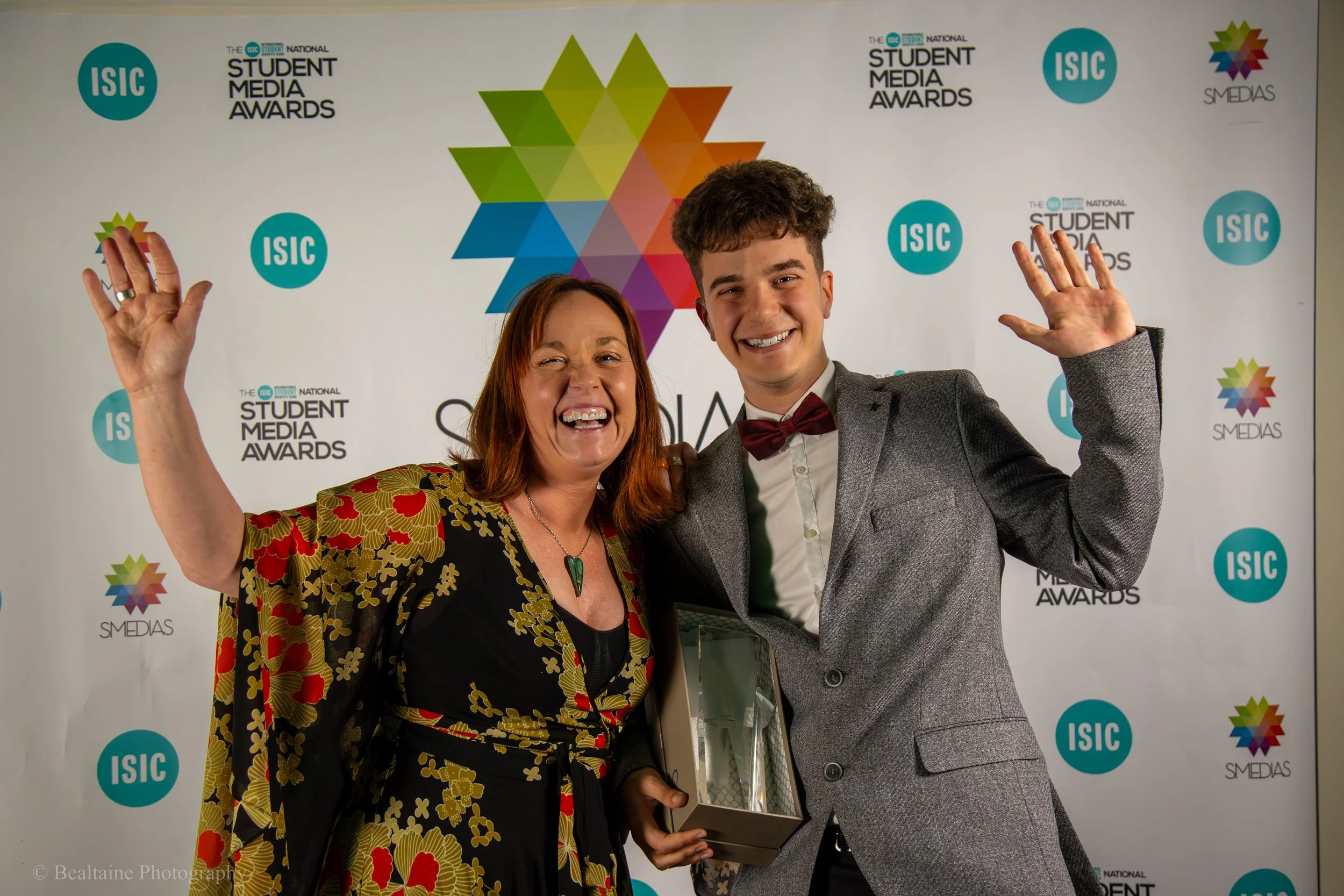 Two people, a woman and a young man, are smiling and waving at an event with a backdrop that reads 'The ISC Student Media Awards.' They are dressed formally; the woman is wearing a patterned dress, and the young man is wearing a suit with a bow tie. 