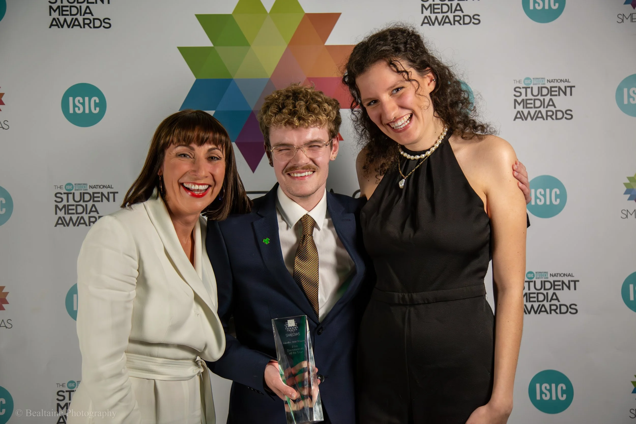 Three people smiling at the camera at the Student Media Awards event, with a colorful geometric background and the event's logo.