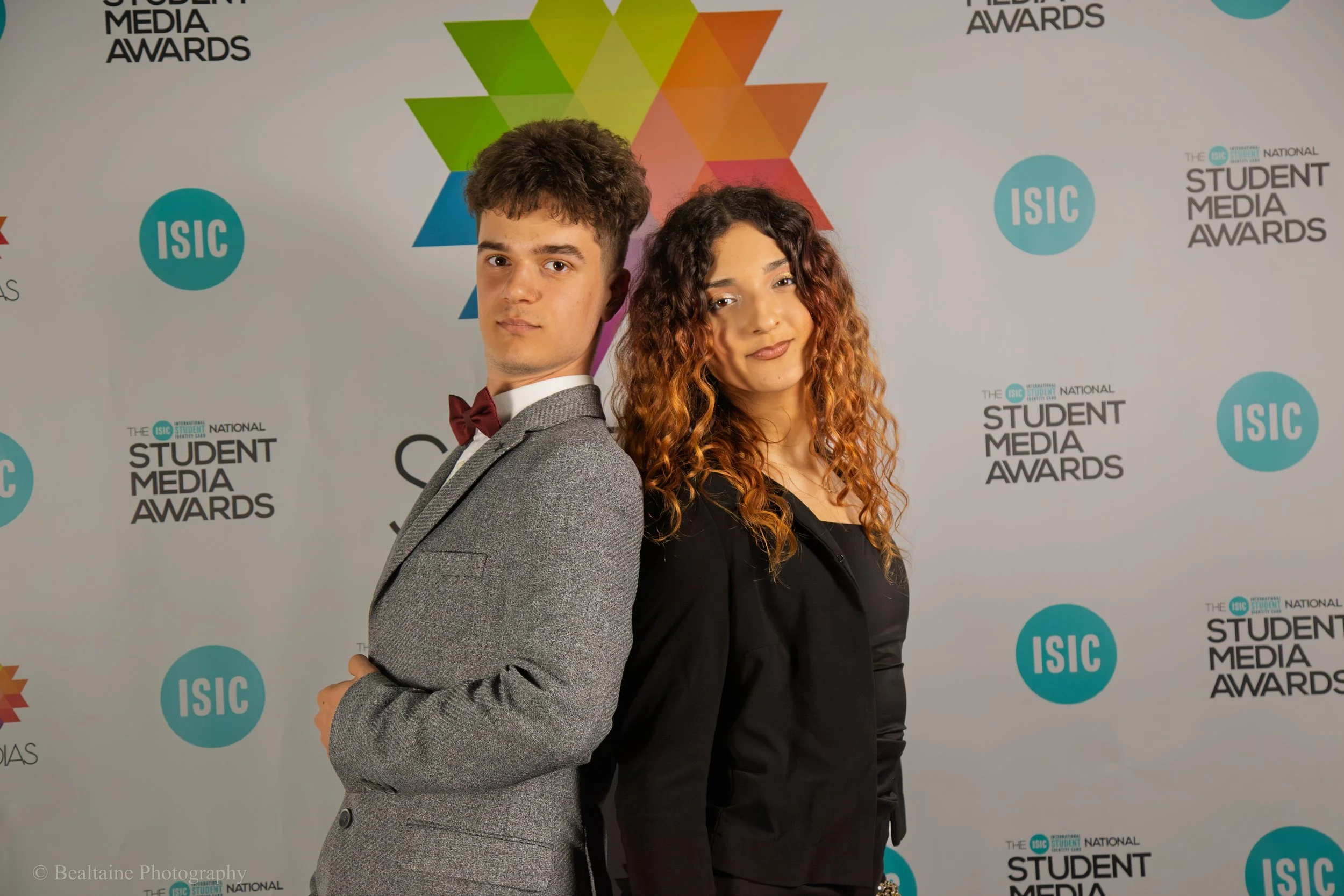 A young man and woman standing back-to-back at the Student Media Awards event with a white backdrop featuring logos and colorful geometric shapes.
