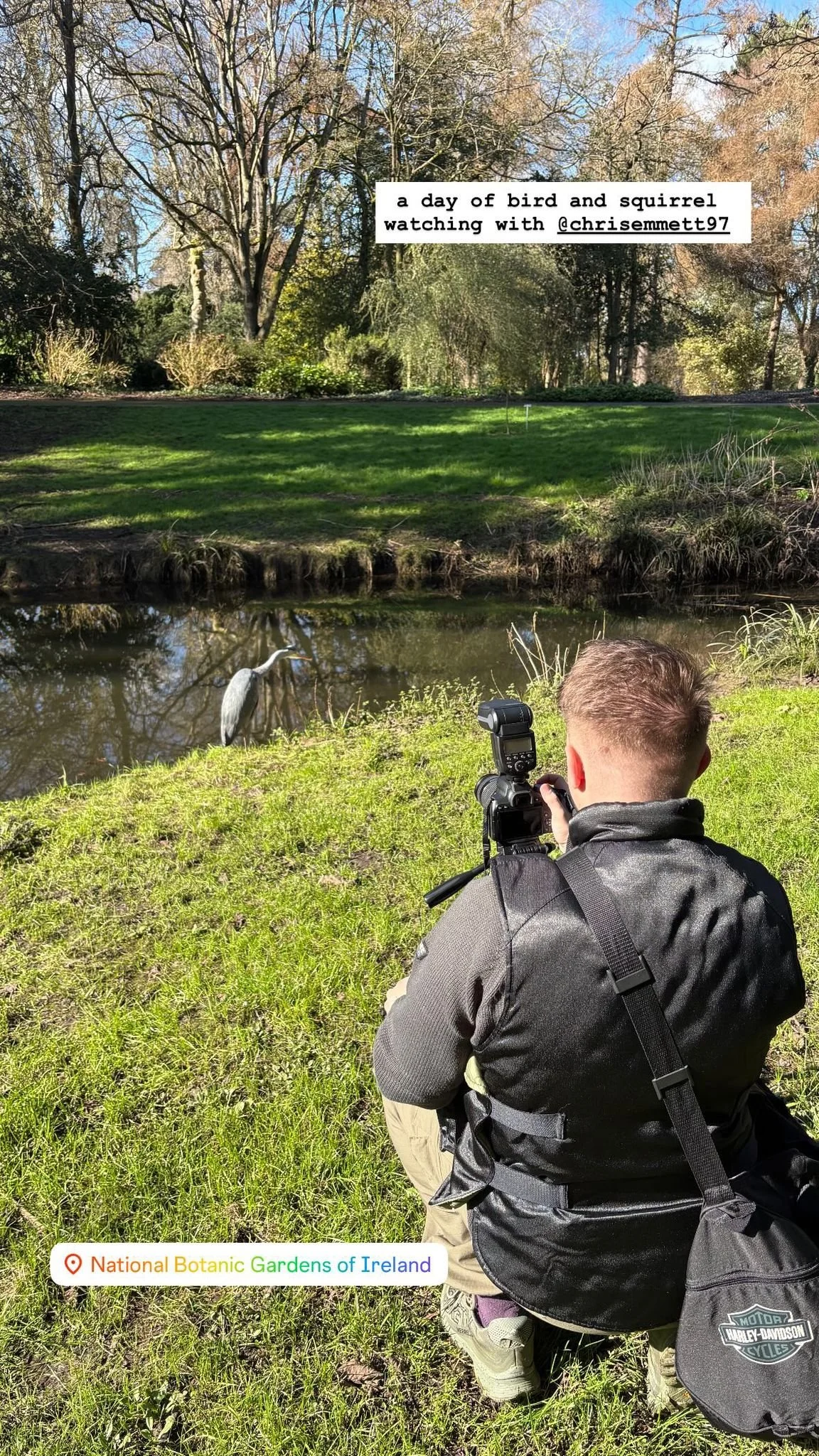 A man with a camera sitting on grass near a pond, watching a grey heron bird standing in the water. The scene is outdoors at the National Botanic Gardens of Ireland on a sunny day with trees in the background.