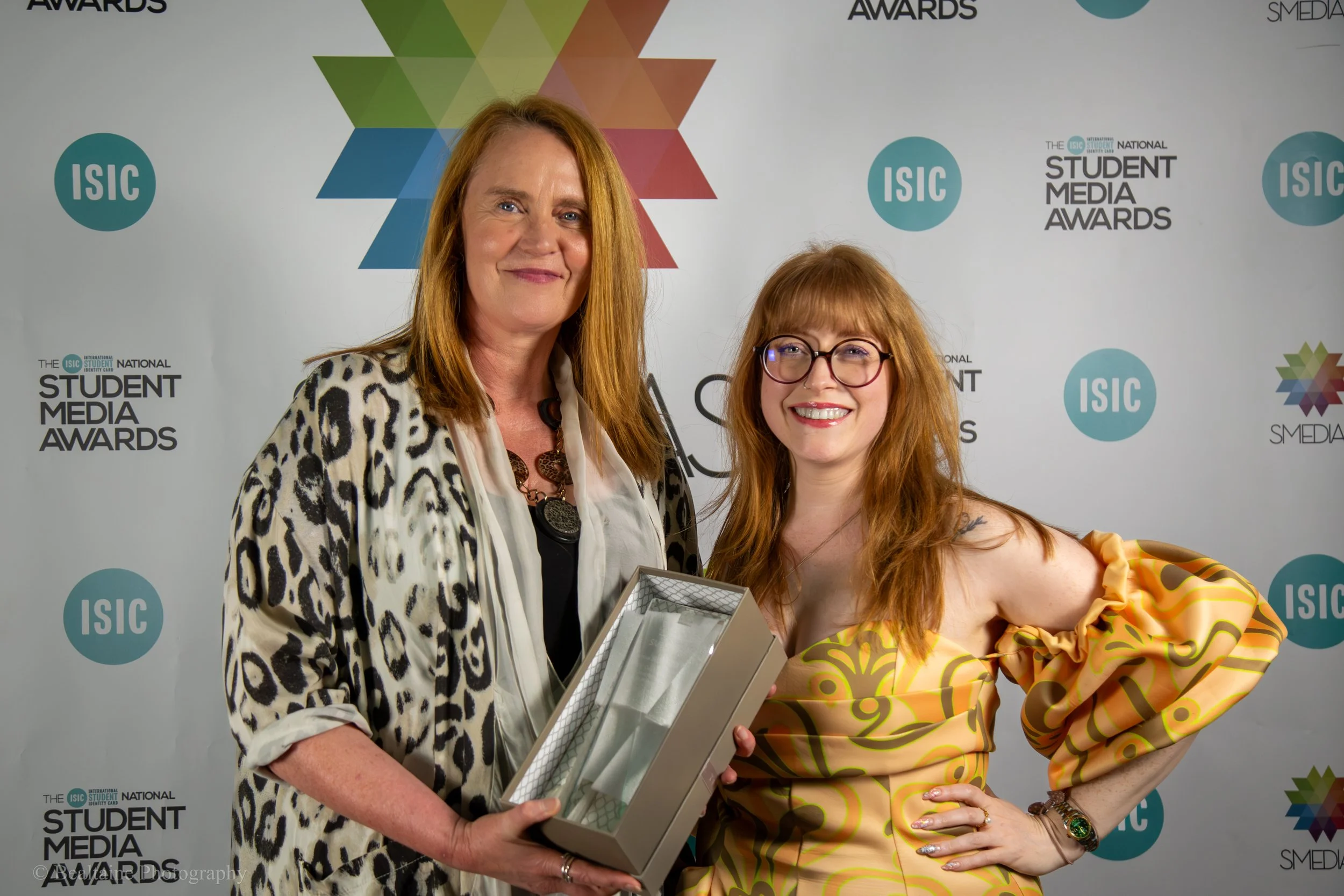 Two women smiling at an awards event, one holding a framed certificate or award. The background features the logos for ISIC and the National Student Media Awards.