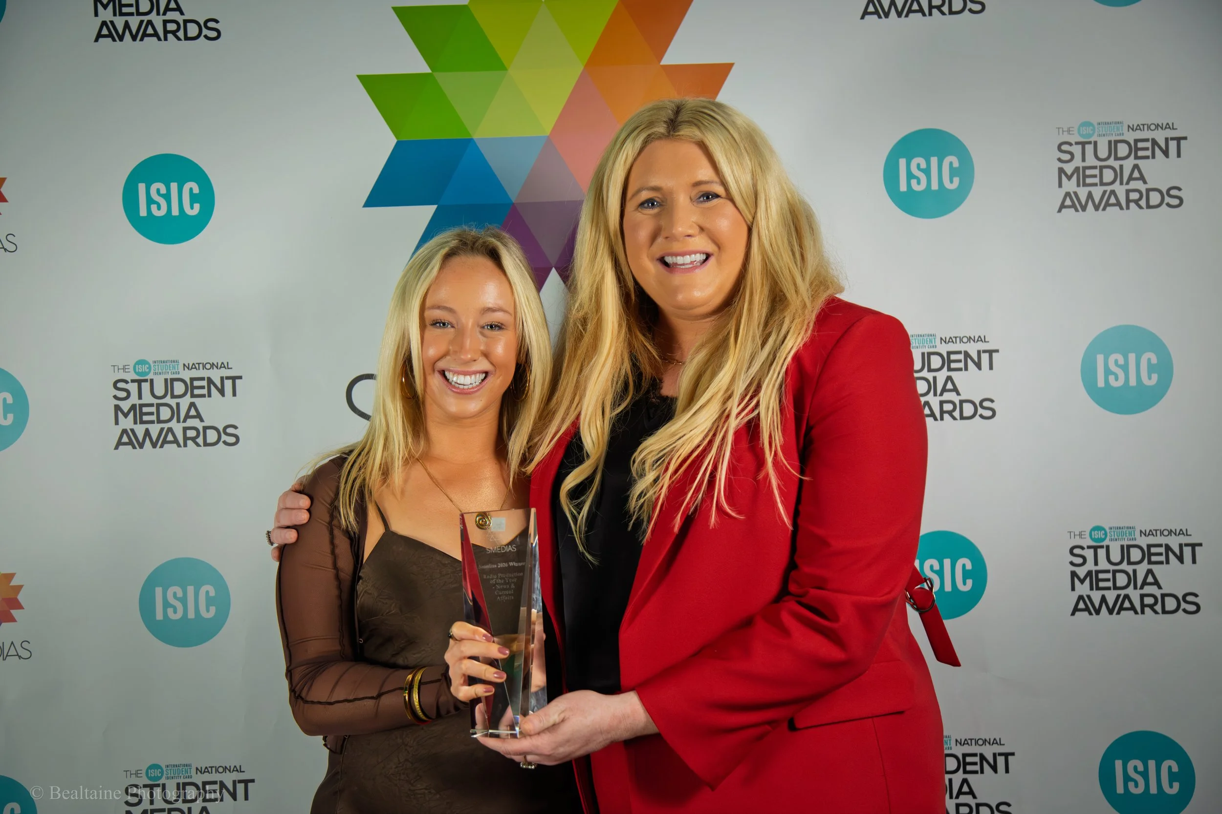 Two women standing together at an awards ceremony, smiling and holding a glass award. One women wears a brown dress, and the other a red blazer. The background features logos and text for the ISC Student Media Awards.