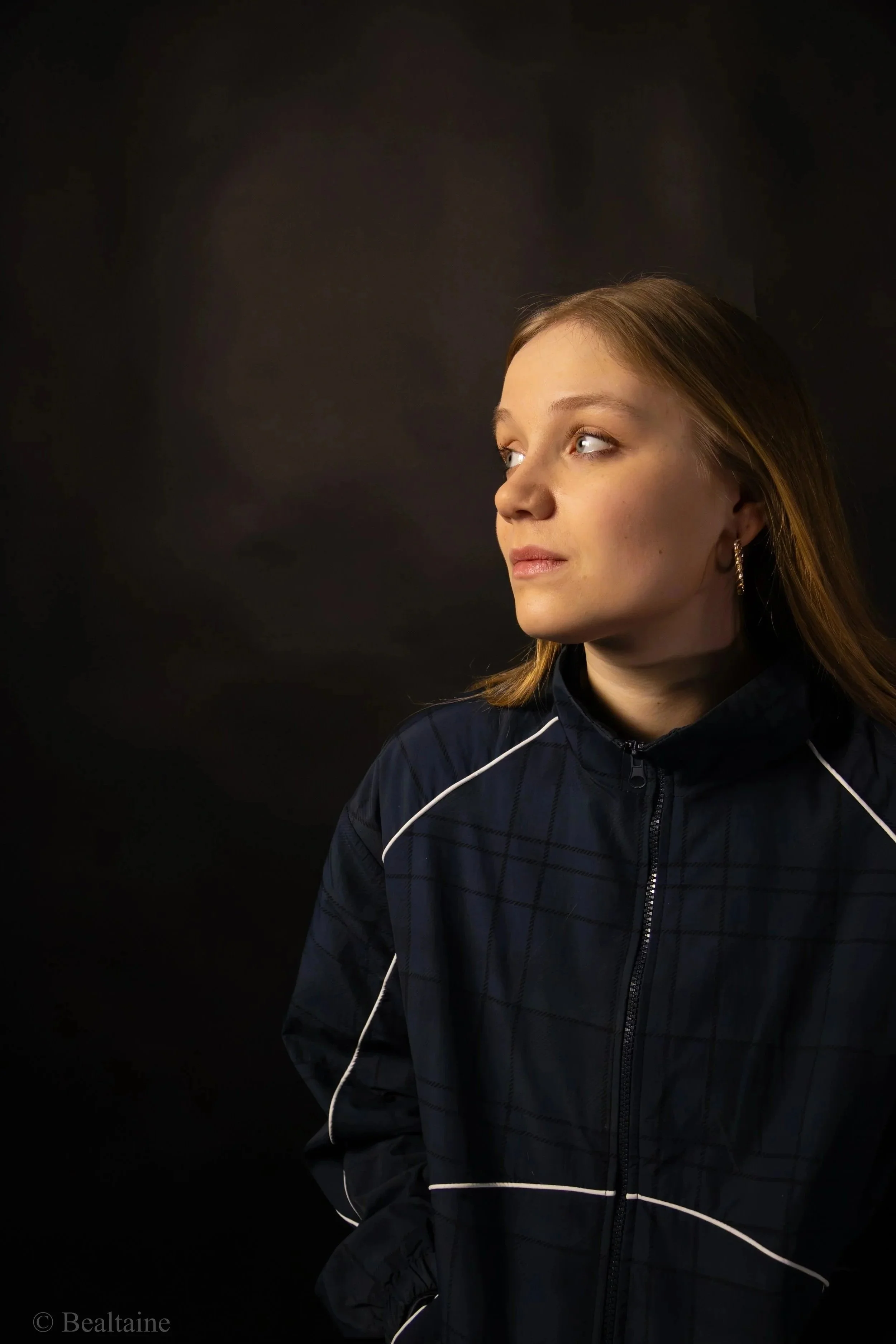 A young woman with brown hair, dressed in a dark sports jacket, looking to her right against a plain black background.