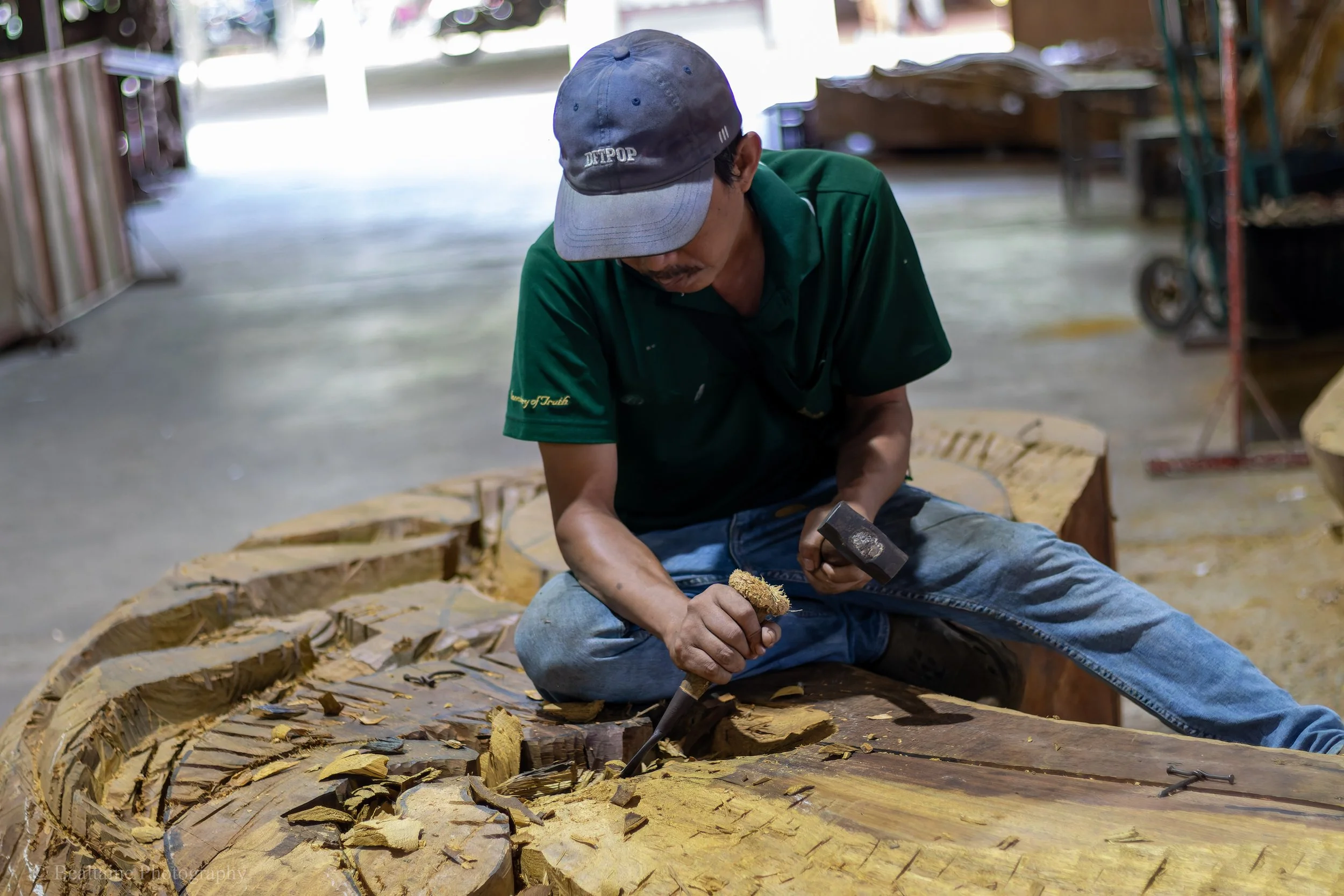 A man wearing a gray cap and green shirt is sitting on the ground, carving or sanding a large wooden boat or log surrounded by wood shavings in a workshop or outdoor setting.