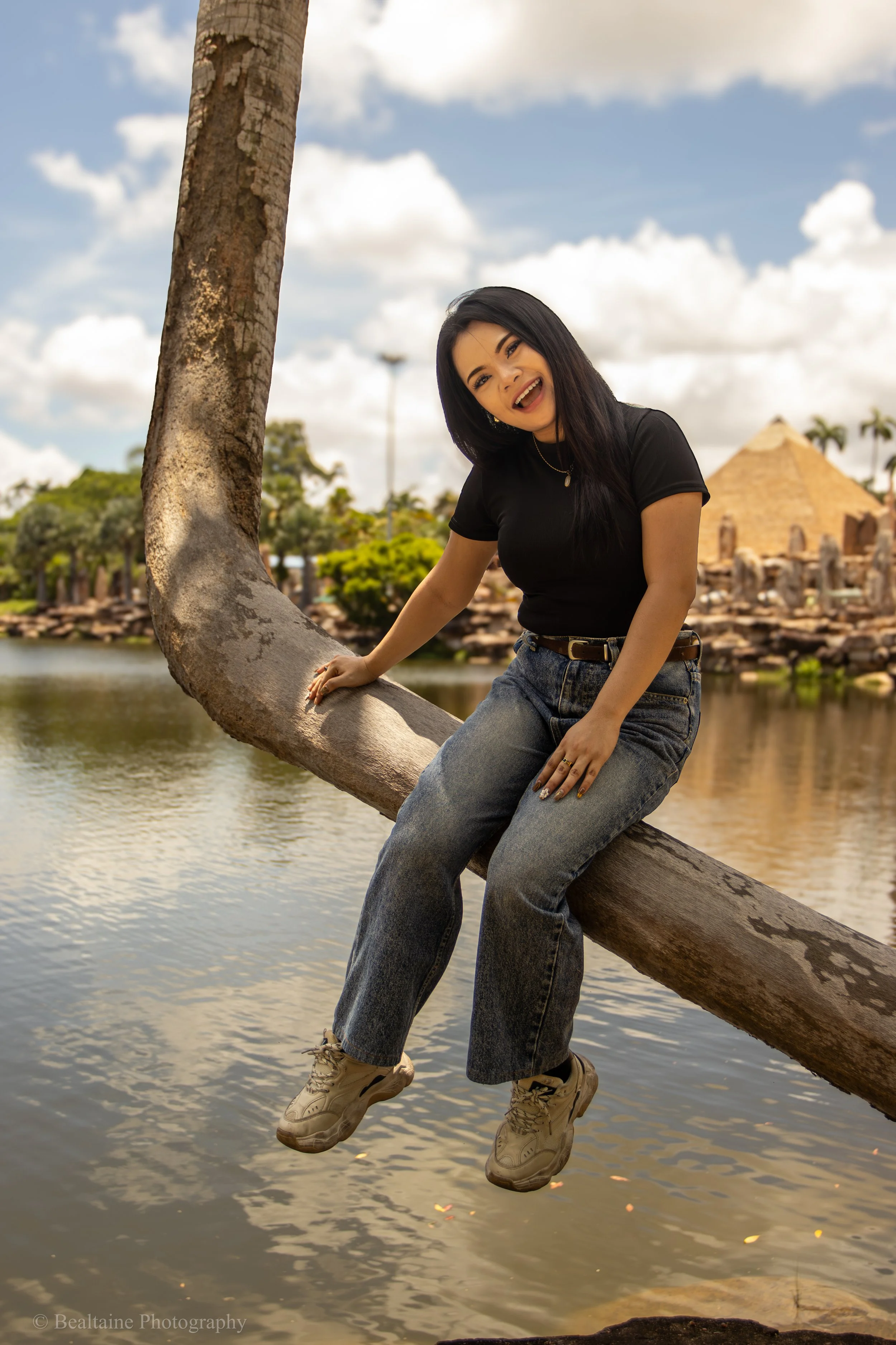 A woman with black hair, wearing a black t-shirt, blue jeans, and beige stylish sneakers, sitting on a large tree branch over a lake, smiling at the camera with a background of a historic building and partly cloudy sky.