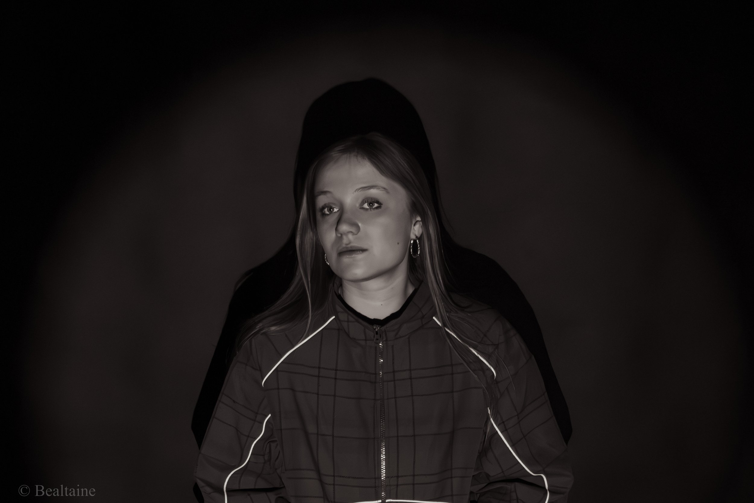 A woman with long hair wearing a dark jacket stands against a dark background. Her face is illuminated, and she has earphones in her ears.