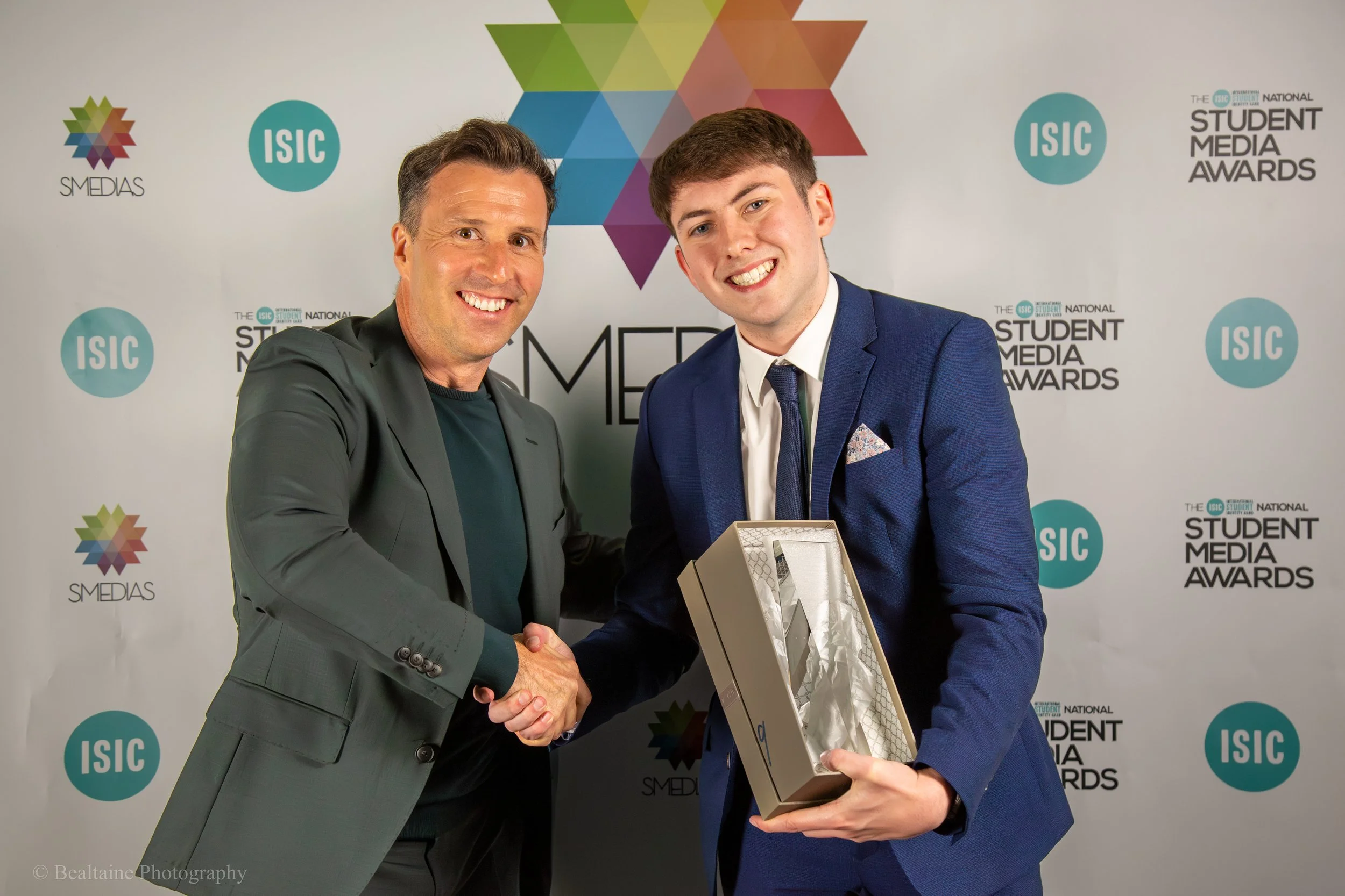 Two men smiling and shaking hands at the Student Media Awards event, with one of them holding a plaque or award. The background features logos and text related to the event.