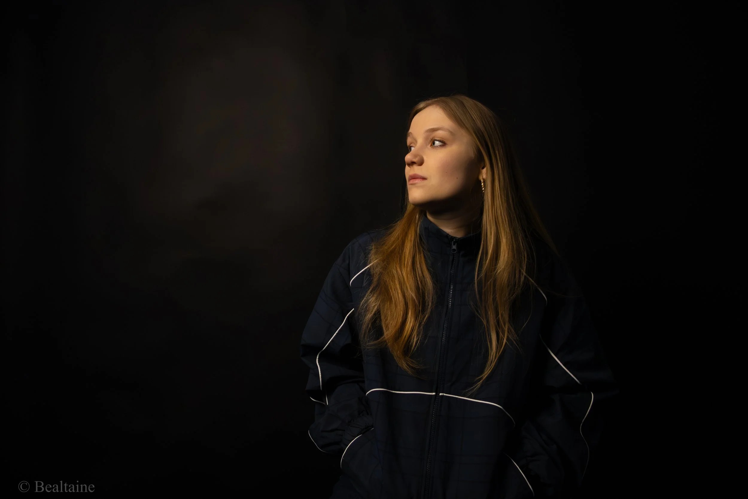 A young woman with long reddish hair looking to her left against a dark background, wearing a black jacket with white accents.