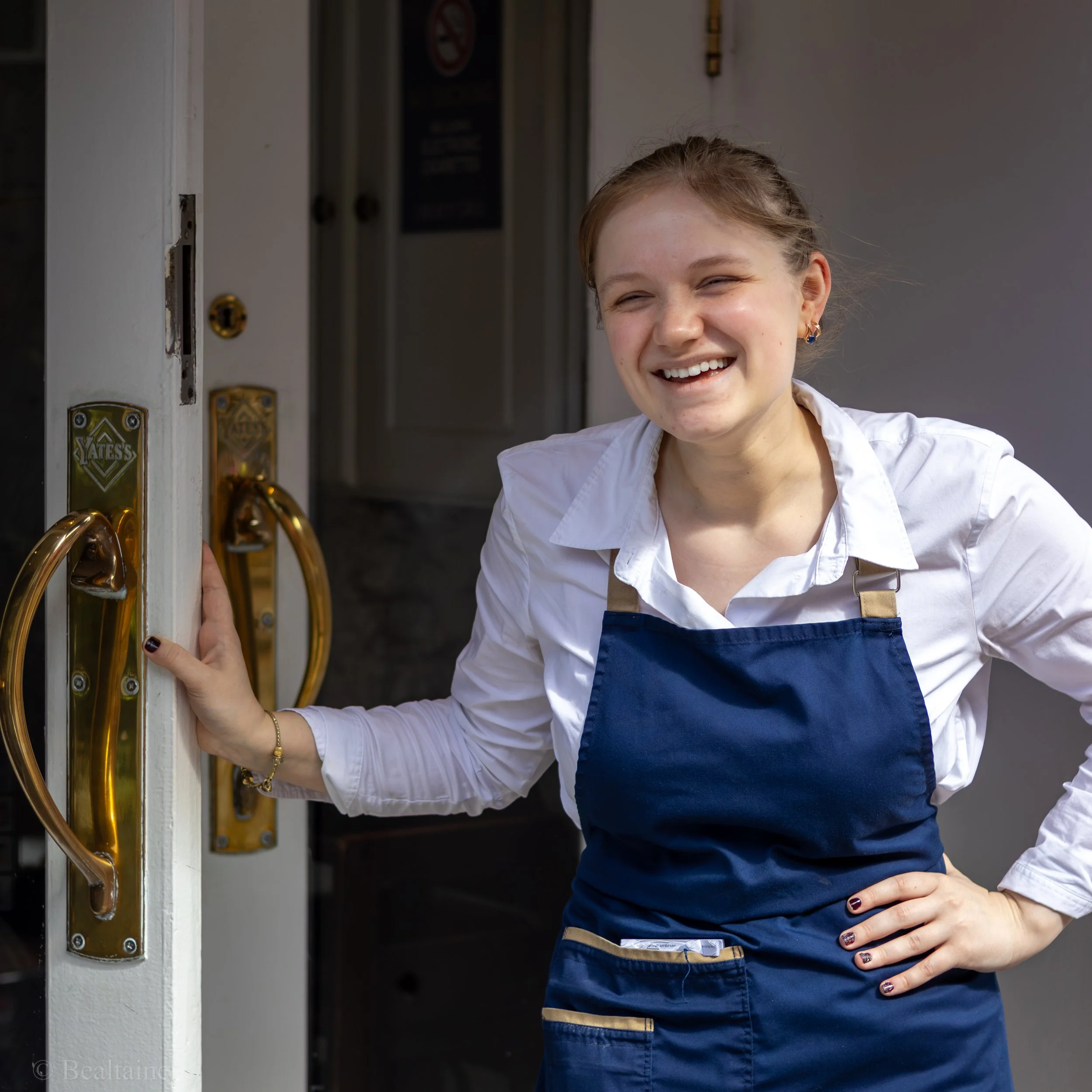A smiling young woman with reddish hair and earrings standing outside a door, holding the door open with her right hand, dressed in a white shirt and a blue apron.