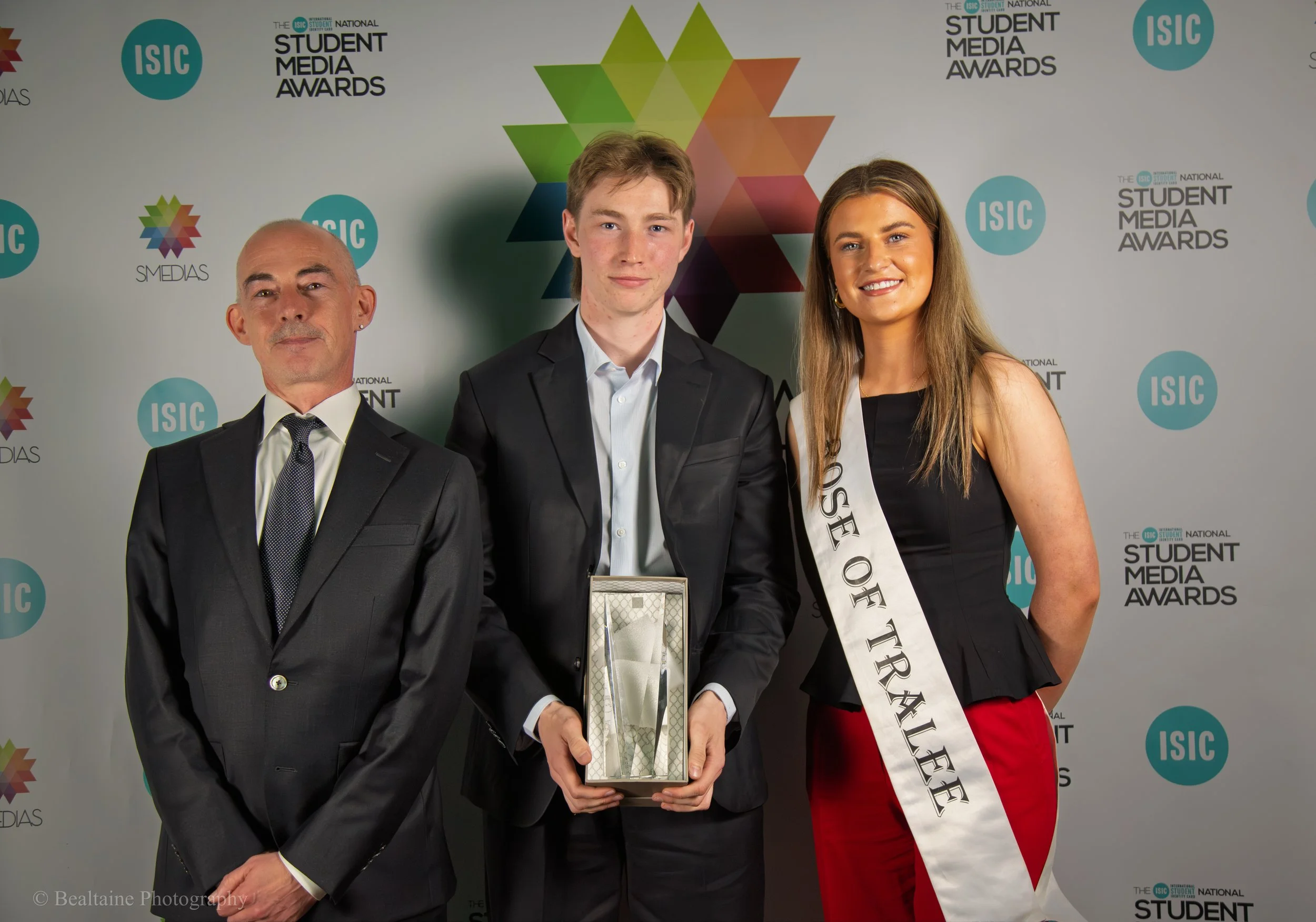 Three people standing in front of a backdrop with logos and text, one man dressed in a black suit and two young adults, one man and one woman, with the woman wearing a sash, all smiling at the camera at the International Student Media Awards event.