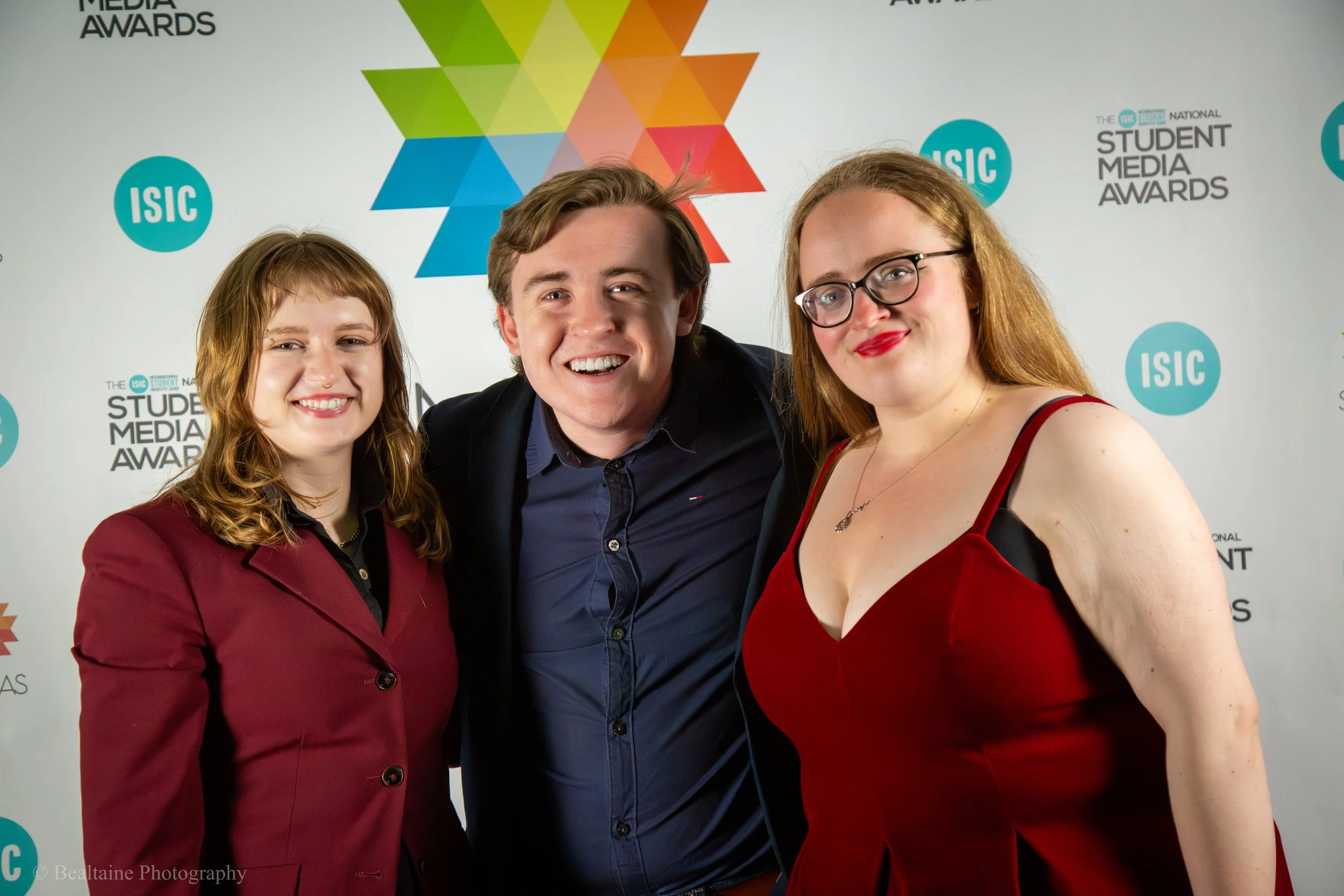 Three people standing together in front of a backdrop with event logos, smiling at the camera. The person on the left has red hair and is wearing a maroon blazer. The person in the middle has light brown hair and is wearing a dark suit jacket. The pe