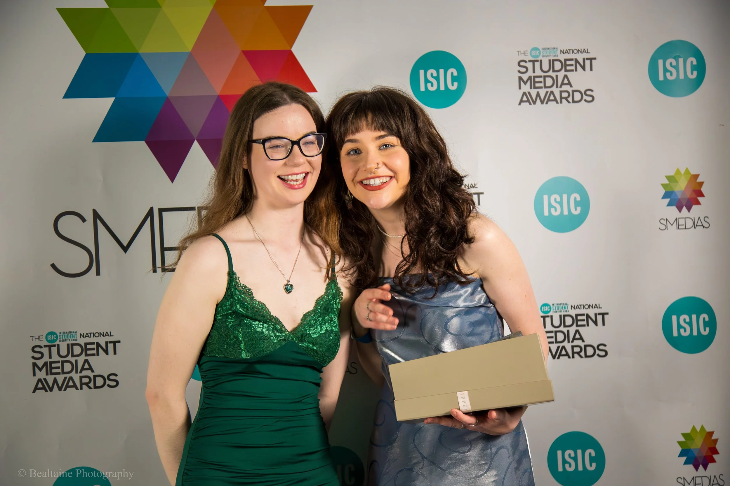Two women smiling at an award ceremony with a backdrop that reads 'Student Media Awards' and features various logos, one of which says 'ISIC.' One woman is wearing a green dress with lace detail and a heart-shaped pendant necklace. The other woman, p