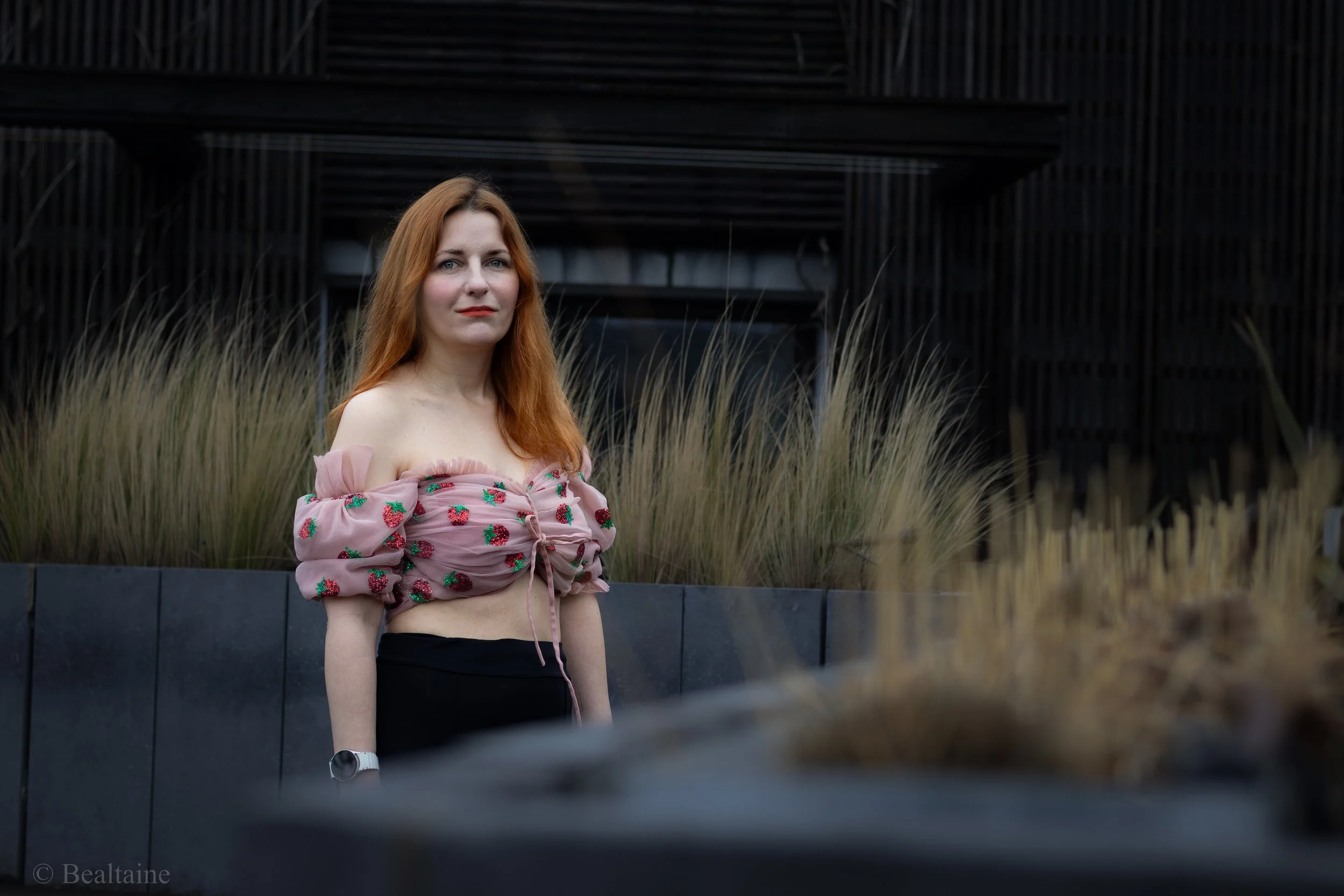 A woman with long red hair and light skin standing outside in front of a modern black building with vertical and horizontal lines. She is wearing a pink off-shoulder top with a strawberry pattern and black pants, looking directly at the camera with a