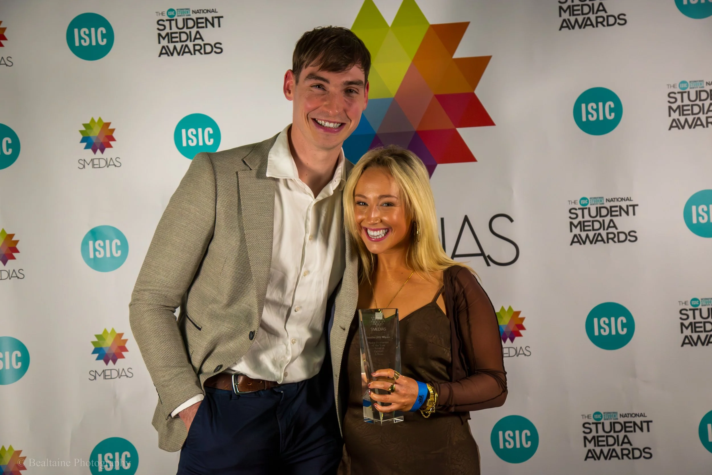 A man and woman standing together at an awards event, smiling. The man is wearing a gray blazer and white shirt, and the woman is dressed in a dark outfit with jewelry. They are posing in front of a backdrop with event logos.