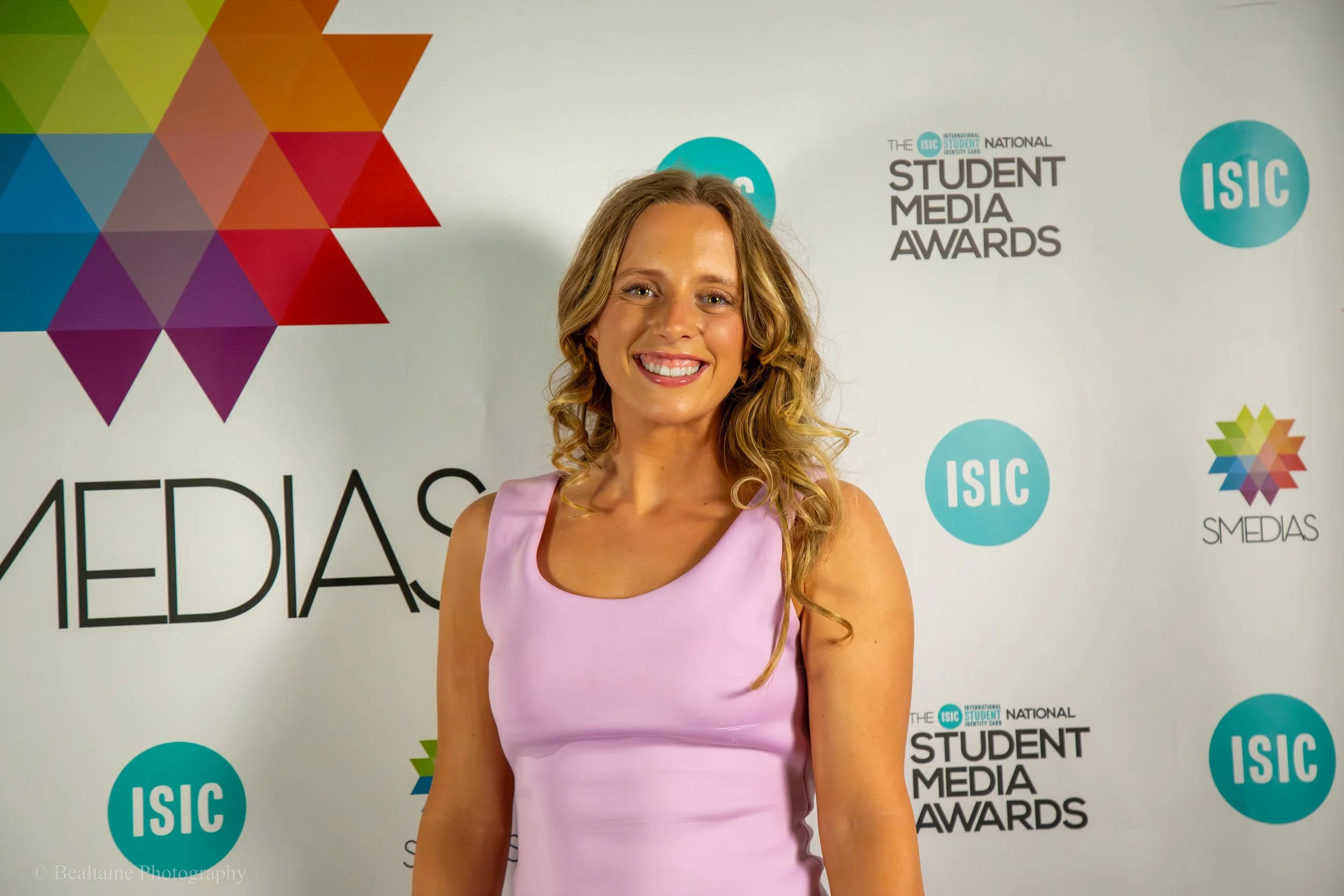 A woman with long curly blonde hair smiling in front of a backdrop with colorful geometric designs and the words 'The ISIC International Student Media Awards' and 'ISIC' logo.