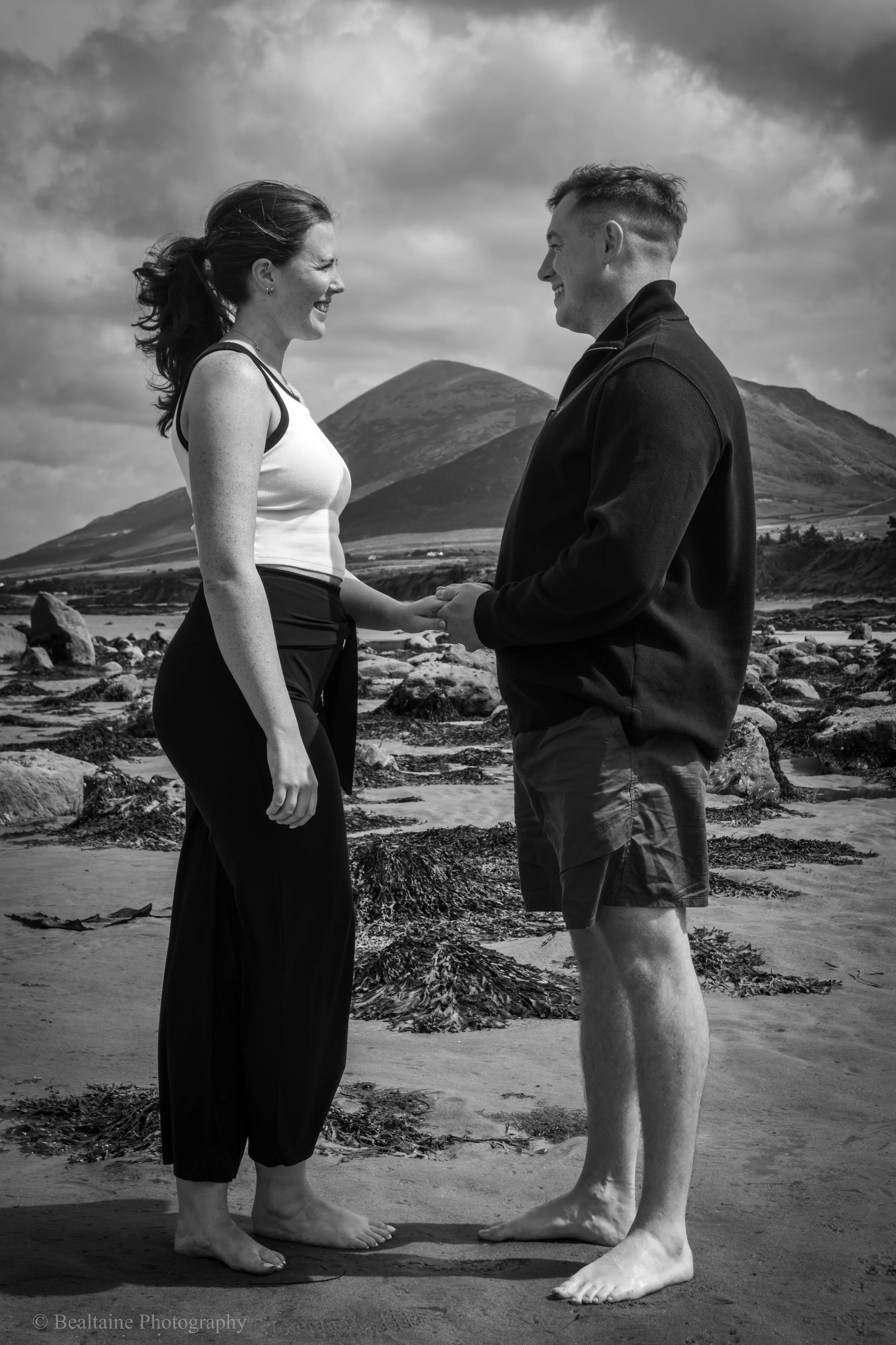 A black-and-white photo of a man and woman standing on a beach, holding hands and smiling at each other, with mountains and clouds in the background.