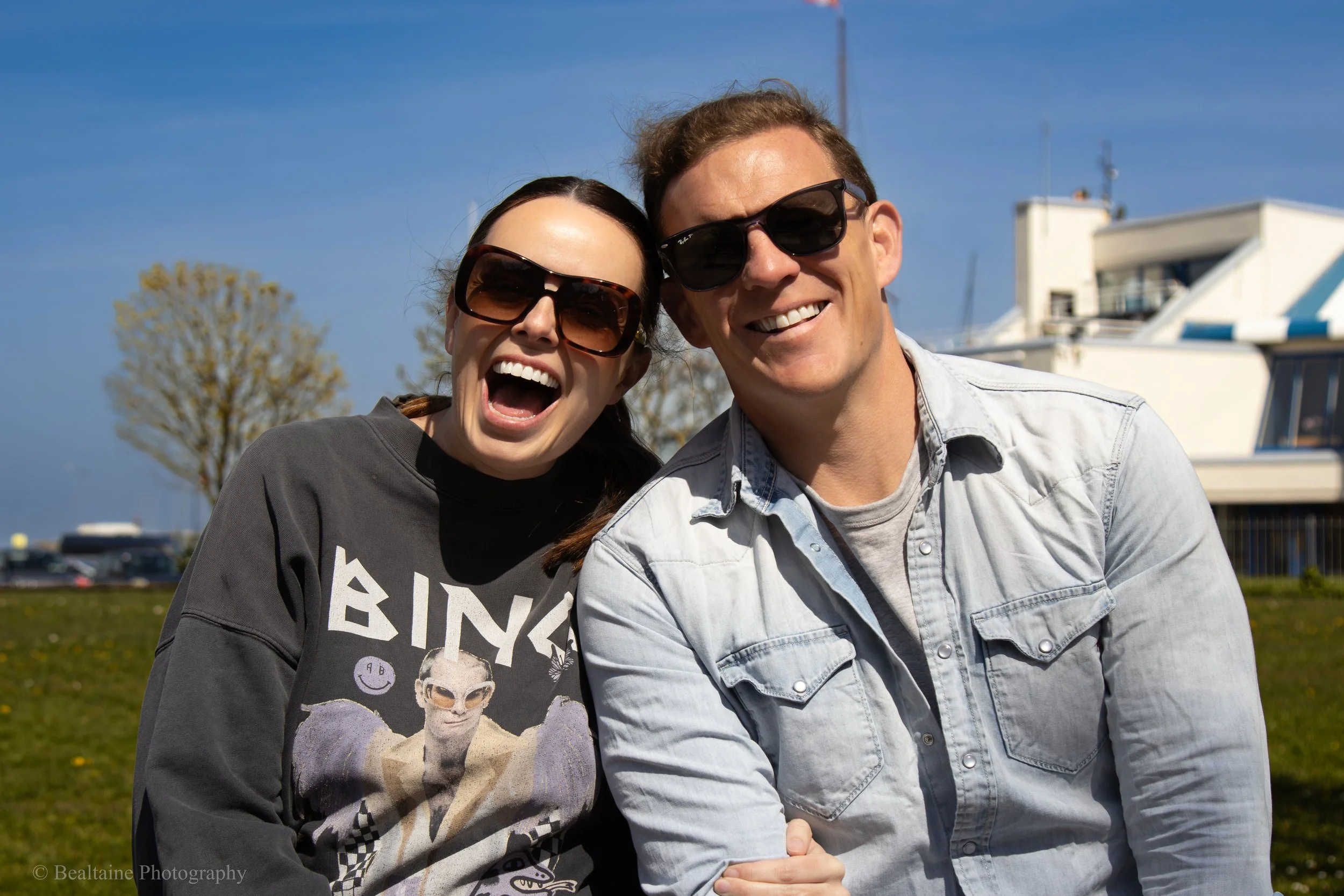 A smiling woman and man wearing sunglasses outdoors on a sunny day. The woman has dark hair and is wearing a dark graphic sweatshirt, and the man has light brown hair and is wearing a light denim jacket. There is a blue sky with some trees and buildi