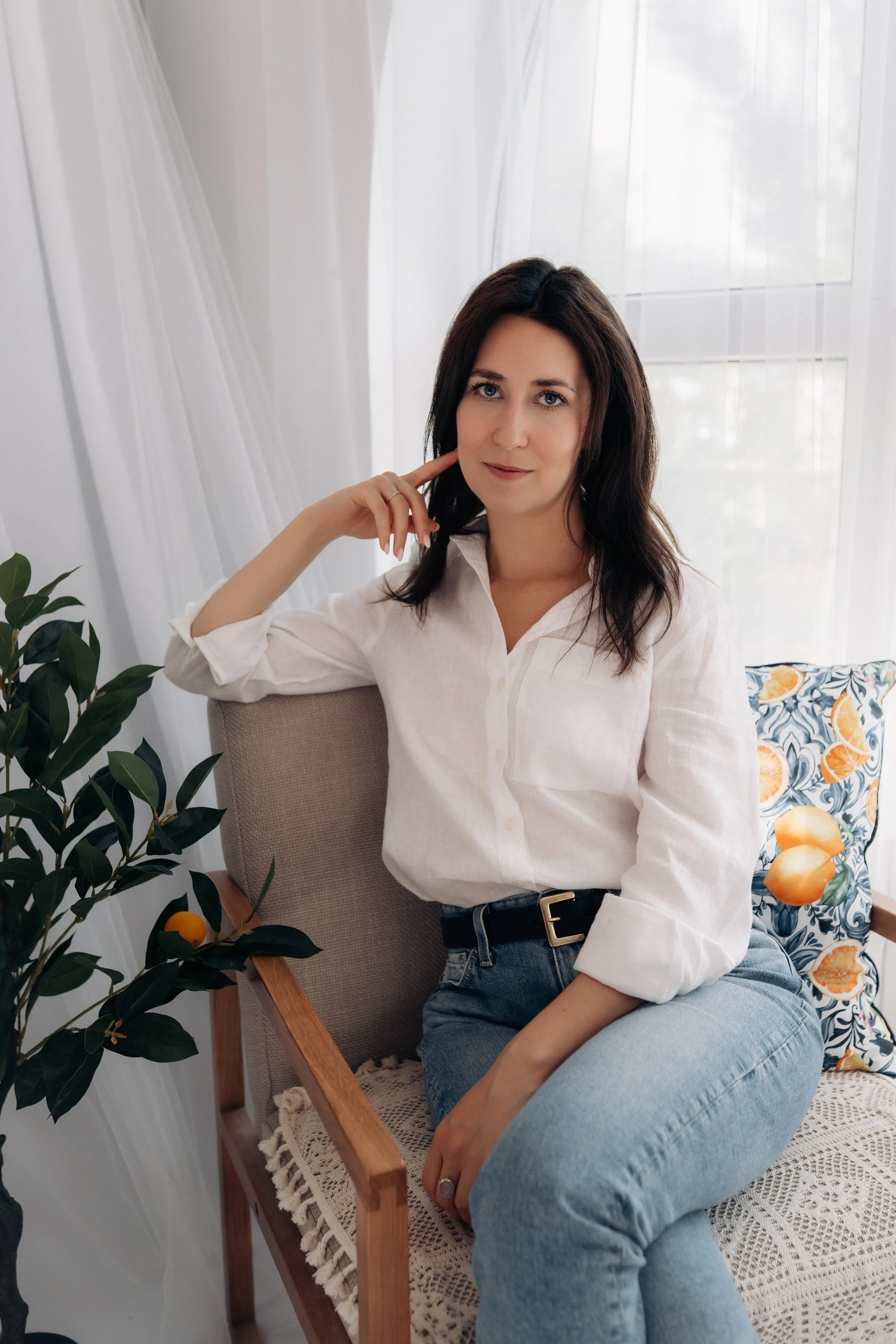 A woman with shoulder-length dark hair, wearing a white button-up shirt and blue jeans, sitting on a wooden chair with a lacy cushion, in a room with white curtains and a window. She is resting her left arm on the armrest and her right hand near her cheek, looking at the camera.