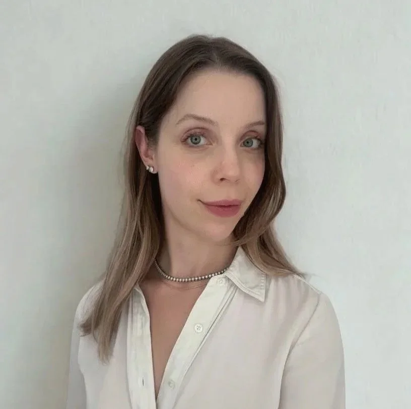 A young woman with shoulder-length light brown hair, wearing a white button-up shirt and pearl jewelry, standing against a plain off-white wall.