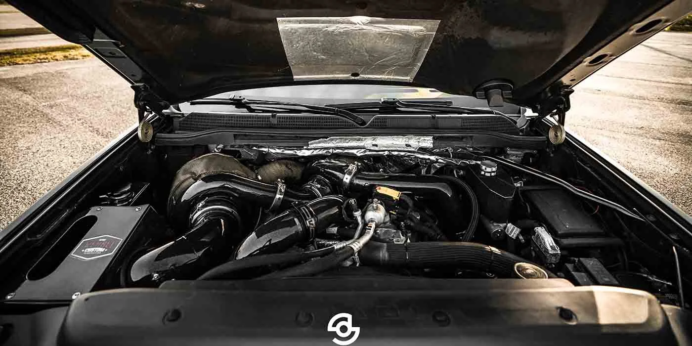 Close-up of a car engine with hood open, showing various engine components and hoses.