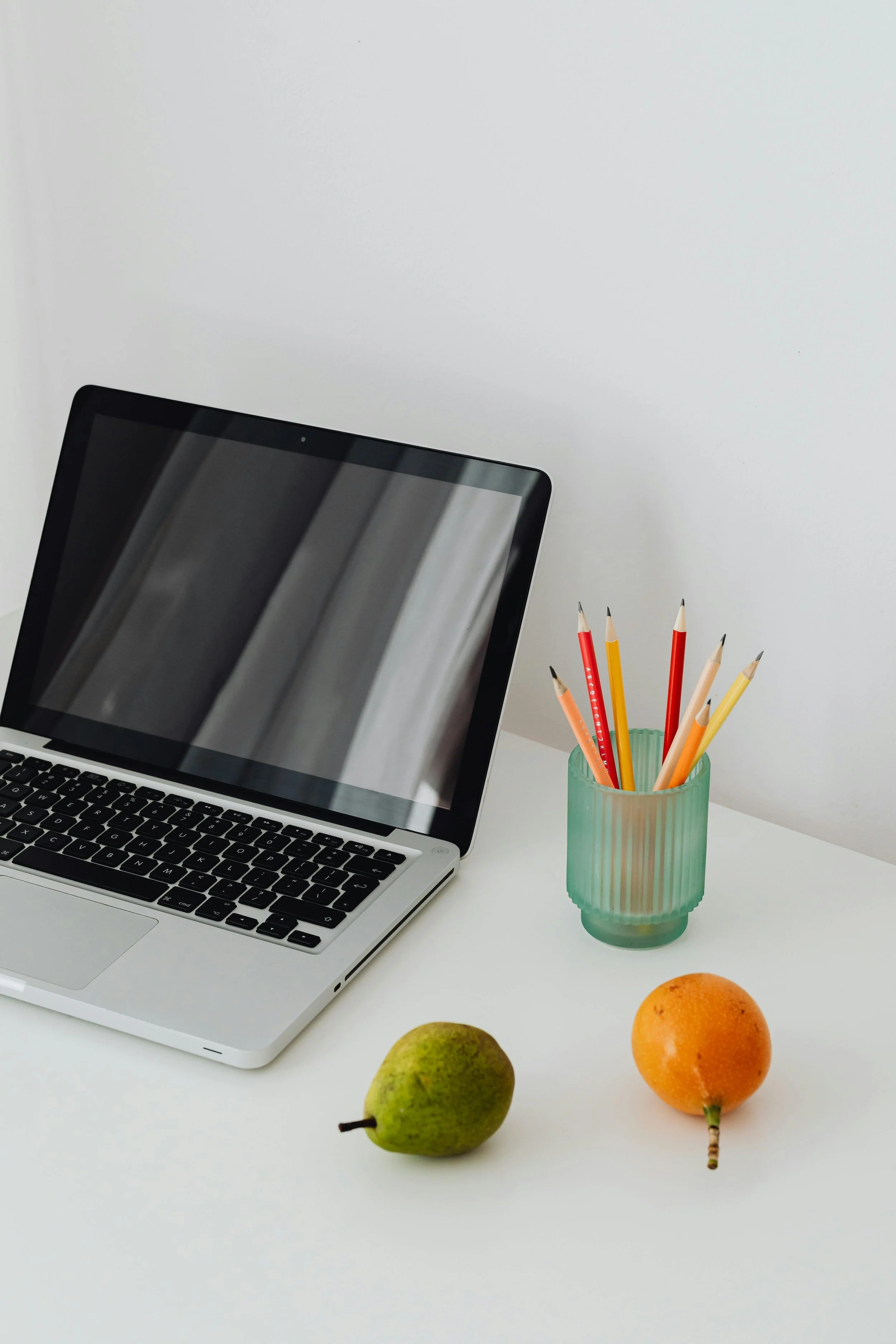 Laptop on a desk with fruit and a glass of colorful pencils