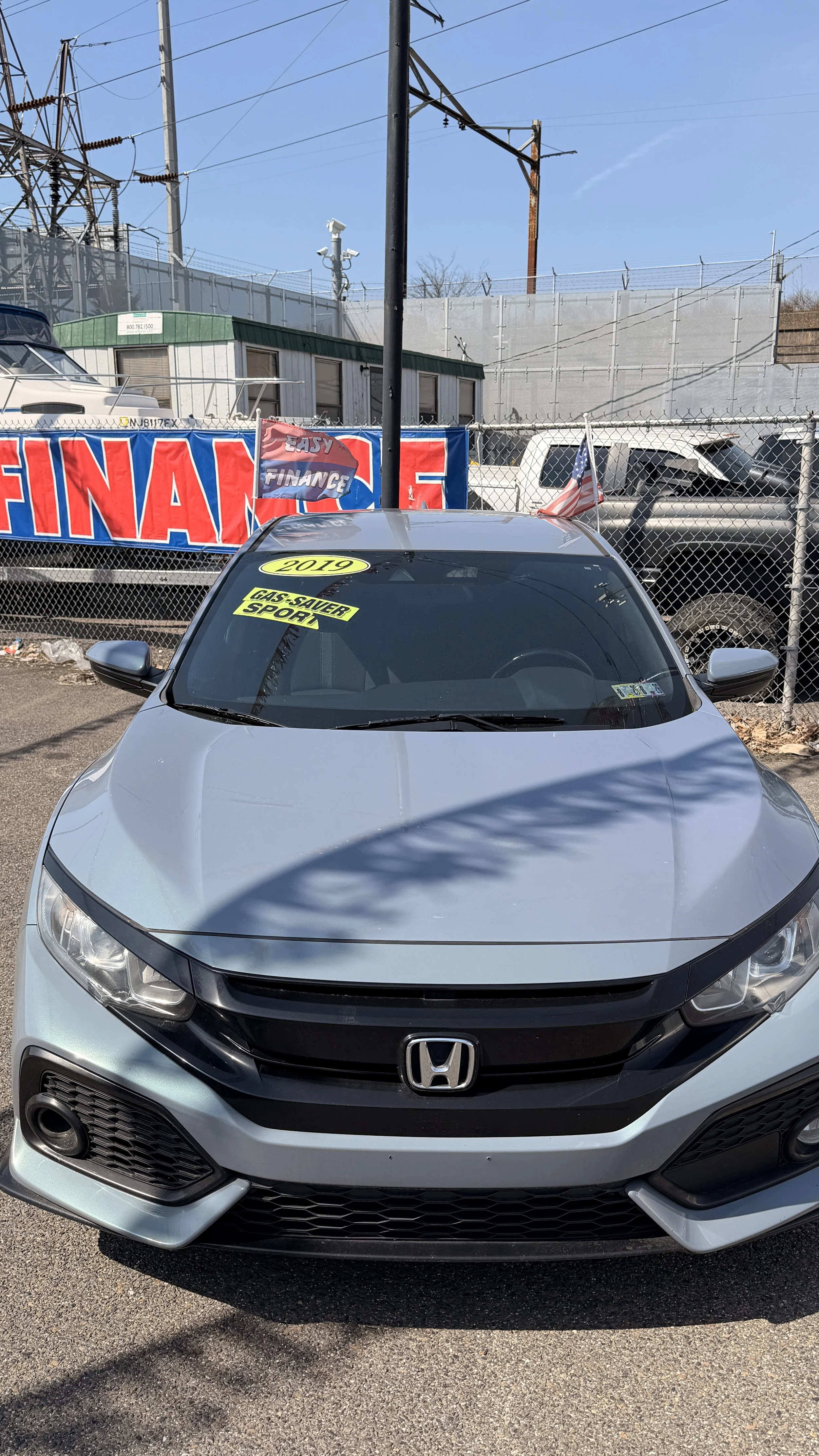 Front view of a silver 2019 Honda sedan parked at a car dealership lot, with yellow tags on the windshield indicating features like 'Gas-Saver' and 'Sport.' A sign in the background says 'Finance' and 'Easy.' The lot is enclosed with a chain-link fence and there are other vehicles and power lines visible at the dealership.