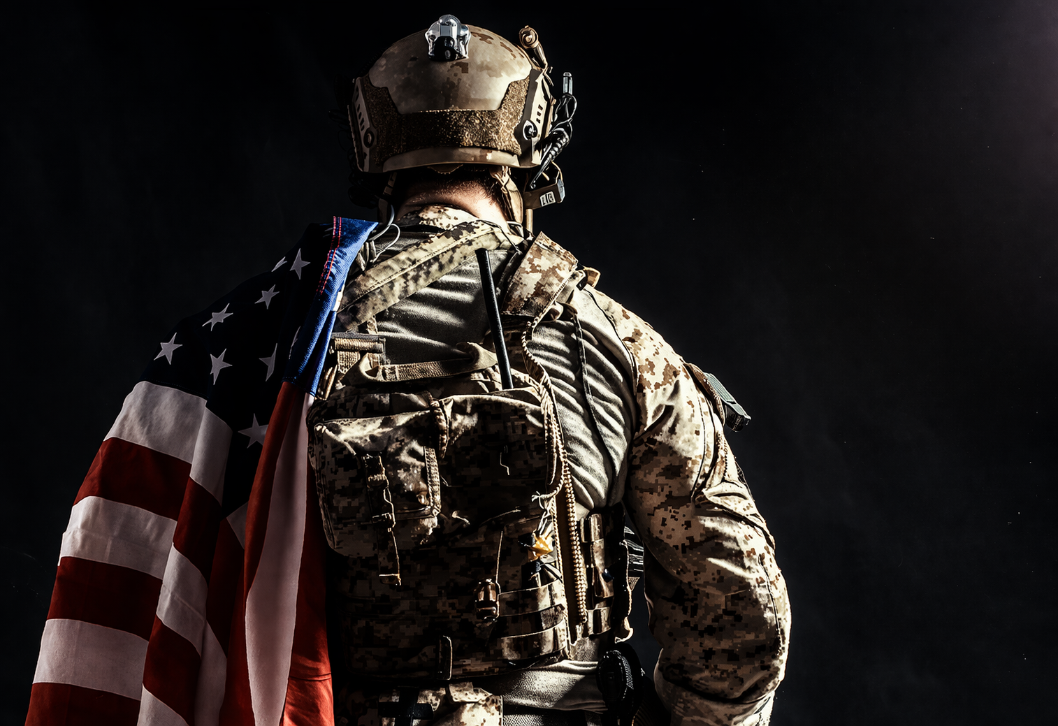 A soldier in full camouflage military gear with an American flag draped over his shoulder, standing against a dark background.