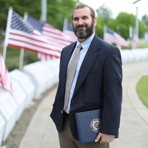 A man with a beard in a suit holding a folder, standing outdoors near a sidewalk lined with American flags.
