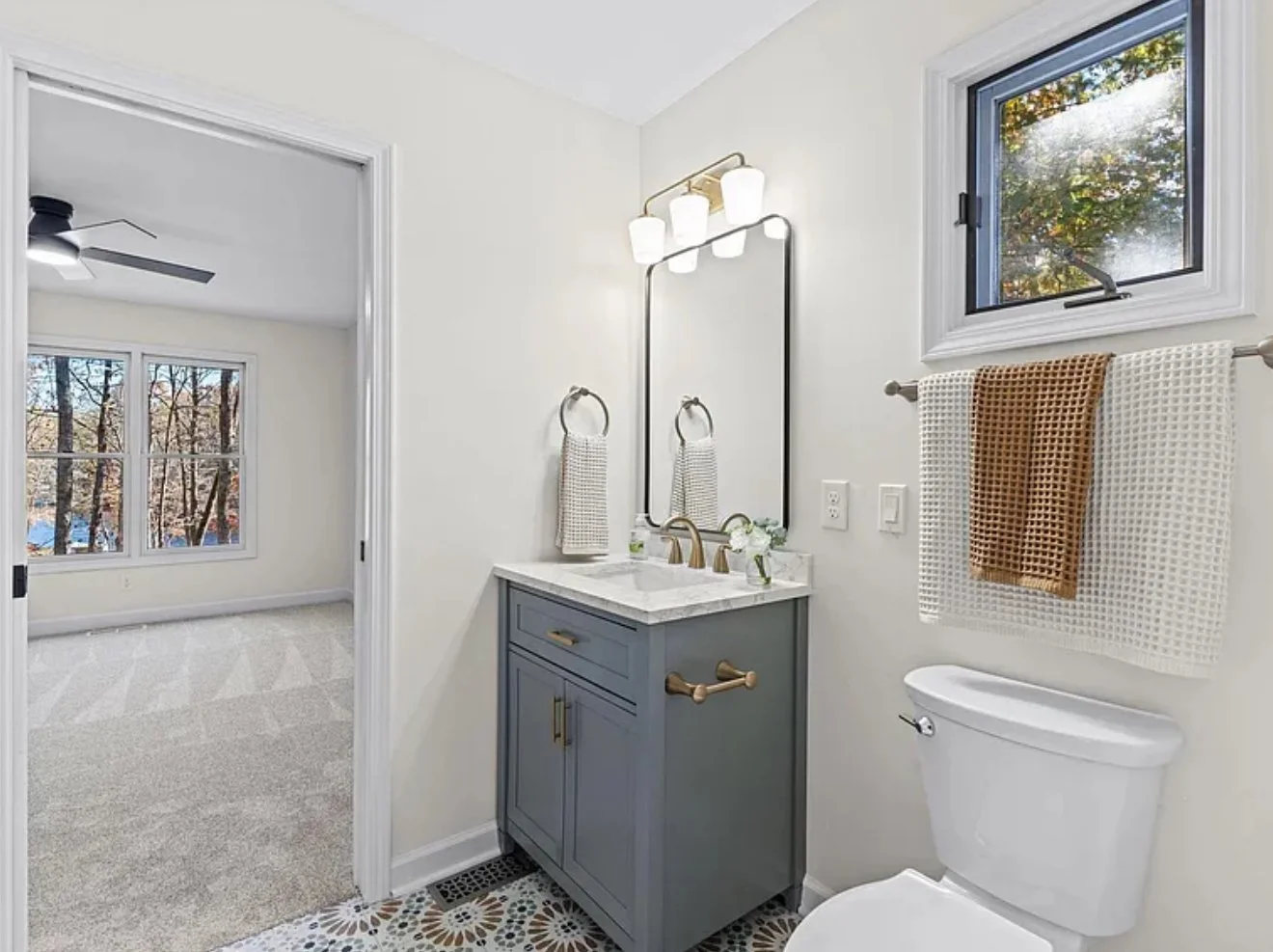 A small bathroom with a gray vanity, white marble countertop, brass fixtures, and a large mirror. There is a towel ring with a striped towel, two decorative towels hanging on a rail, a small window with a view of trees, and a set of three white lights above the mirror.