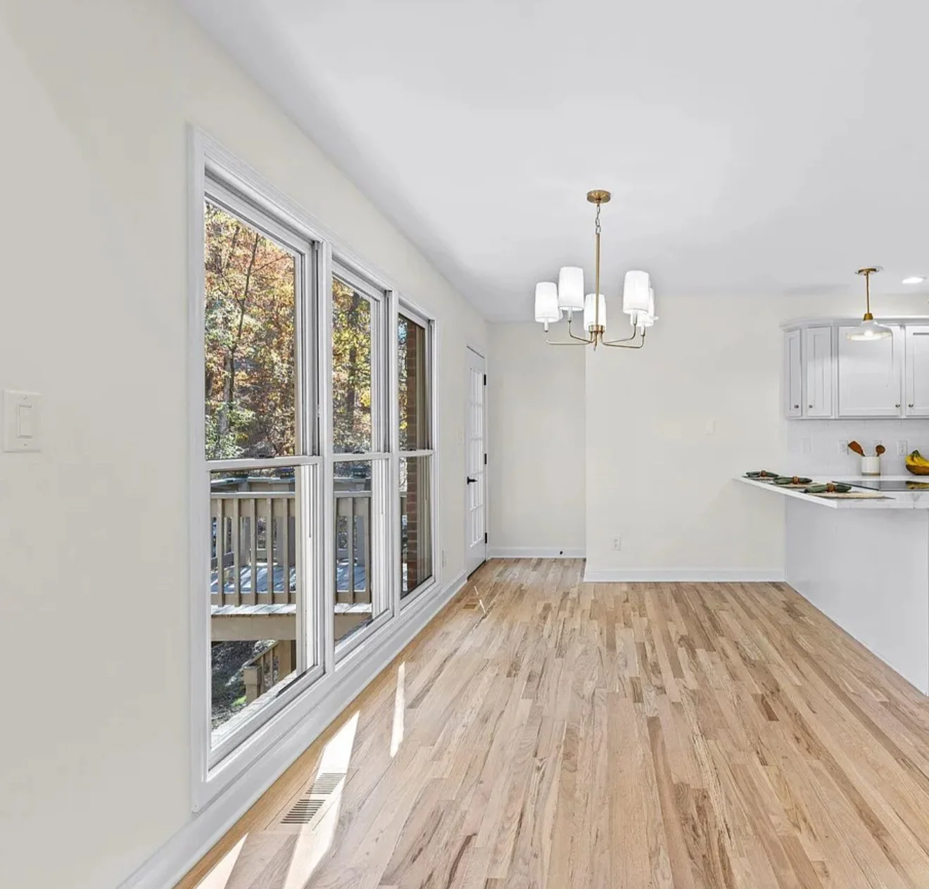 Empty bright living area with large windows, white walls, hardwood floors, and a kitchen in the background with white cabinets and ceiling lights.