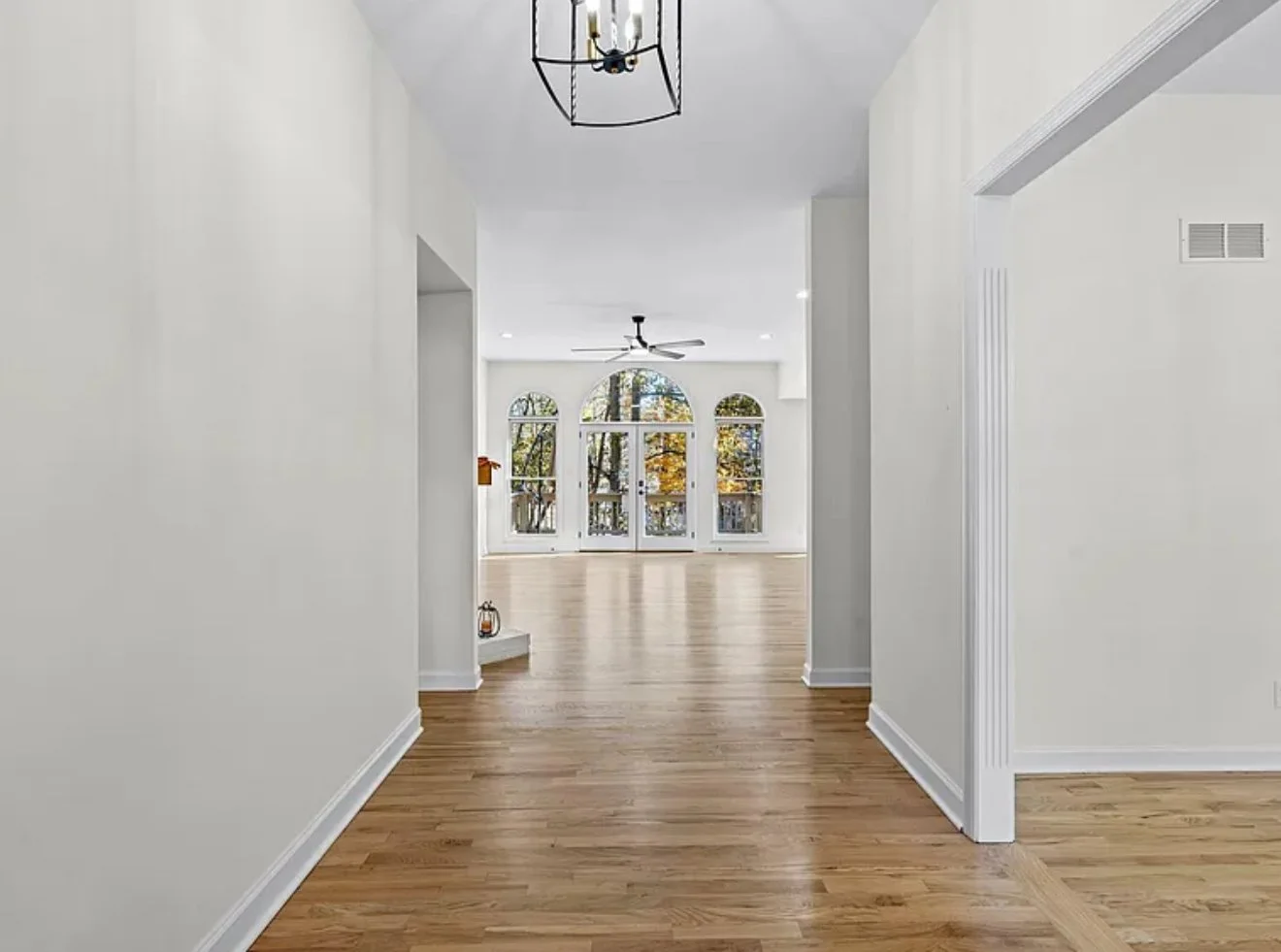 Empty living space with hardwood floors, white walls, large windows with trees outside, ceiling fan, and ceiling light fixture.