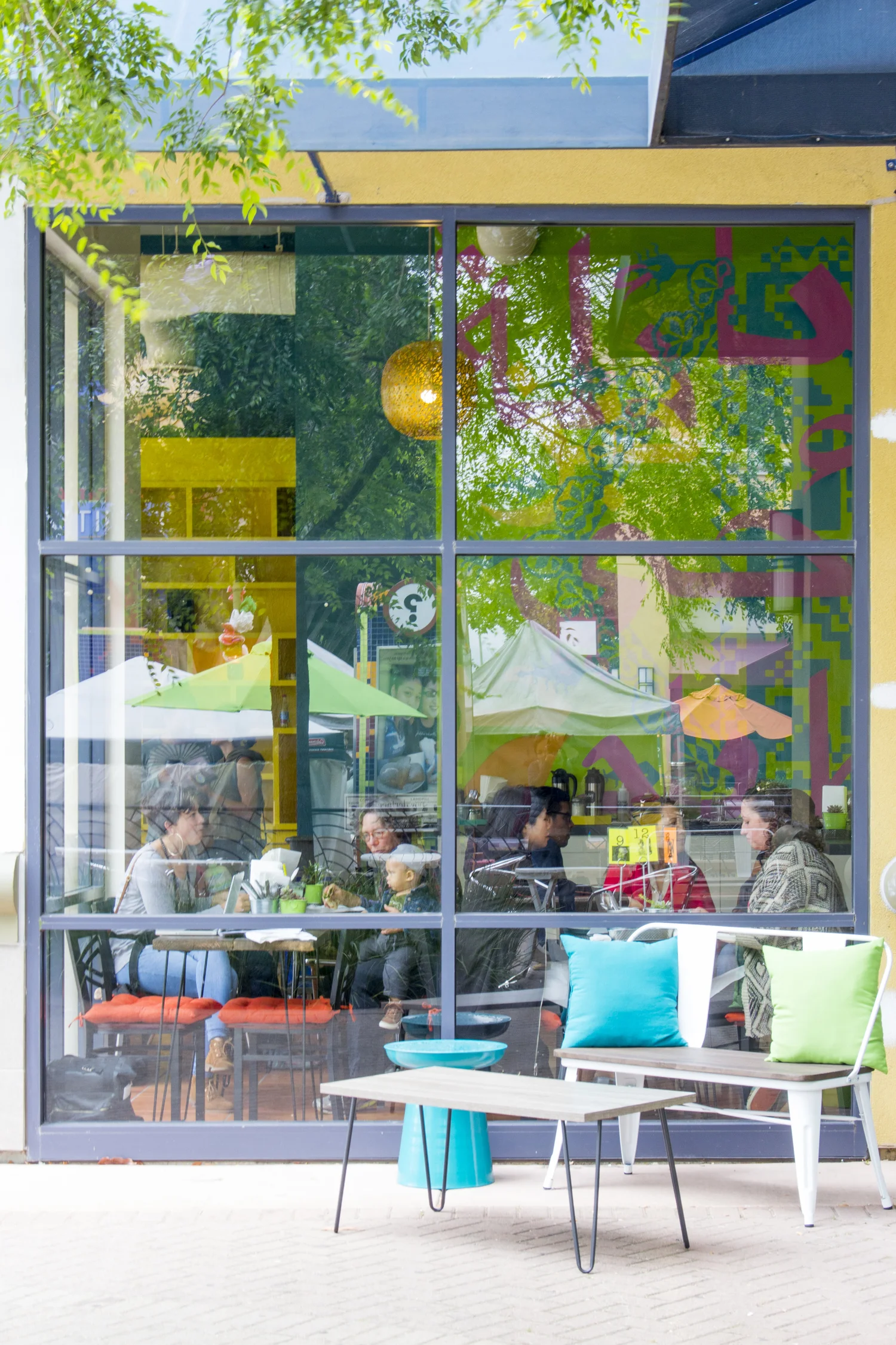 View of a large glass facade, with outdoor seating in the foreground and patrons, including a child on his mother's lap, eating and socializing.