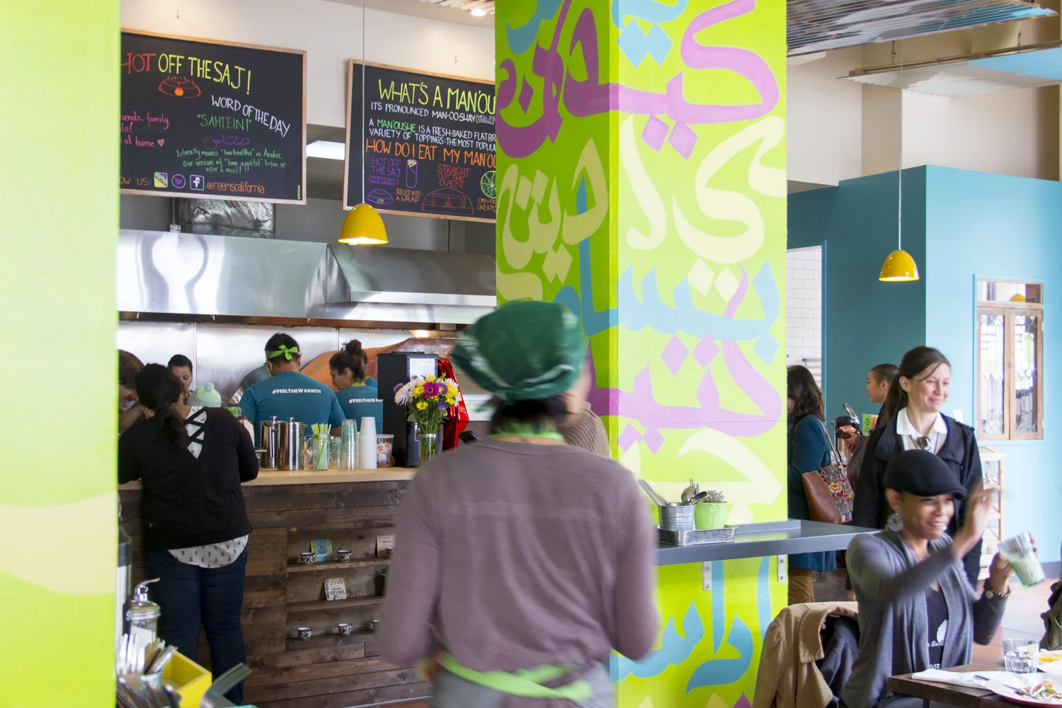 A bustling, brightly painted interior, with chalkboard menu boards and custom mural with Arabic lettering.
