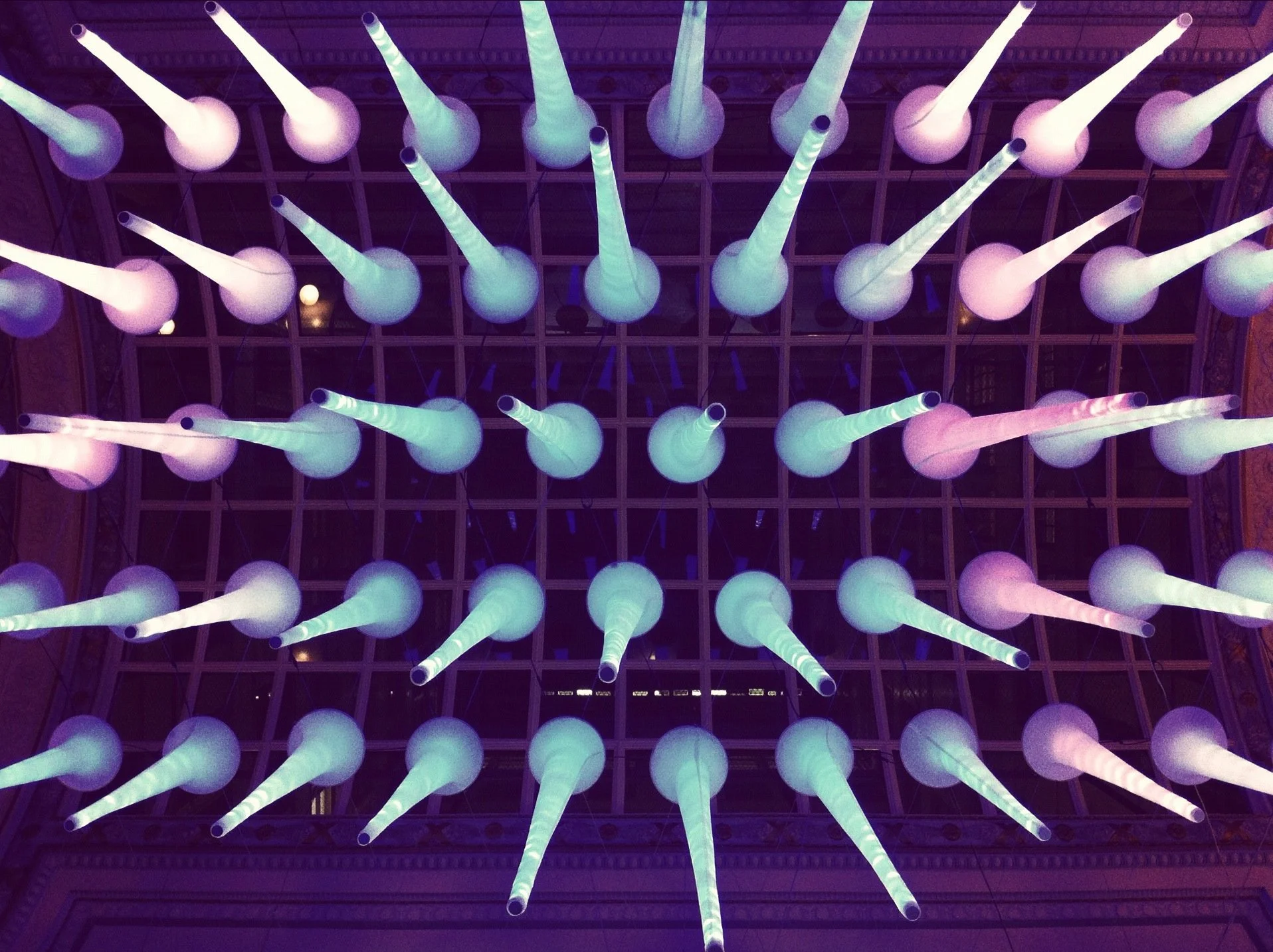 Ceiling installation with multiple large, illuminated, paper or plastic cone-shaped light fixtures hanging from a grid ceiling, emitting soft purple, pink, and greenish light.