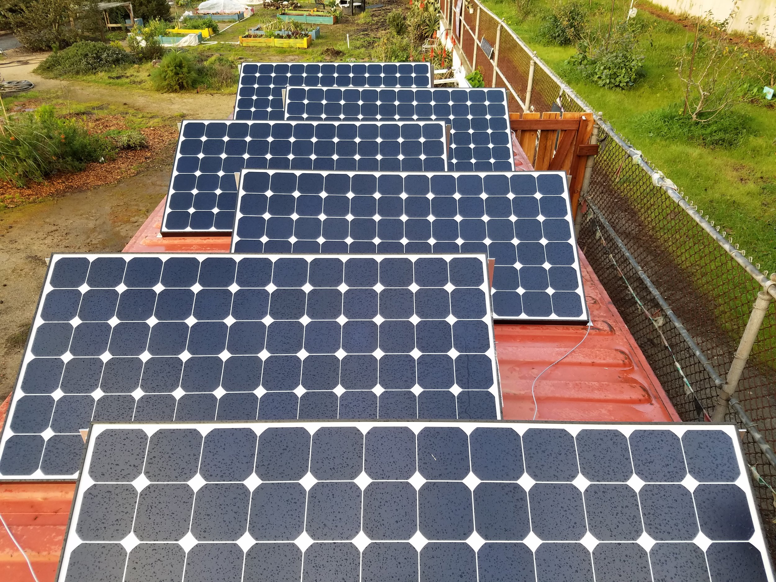 Multiple black solar panels installed on a red flat roof of a building, with a garden and raised planting beds visible in the background.