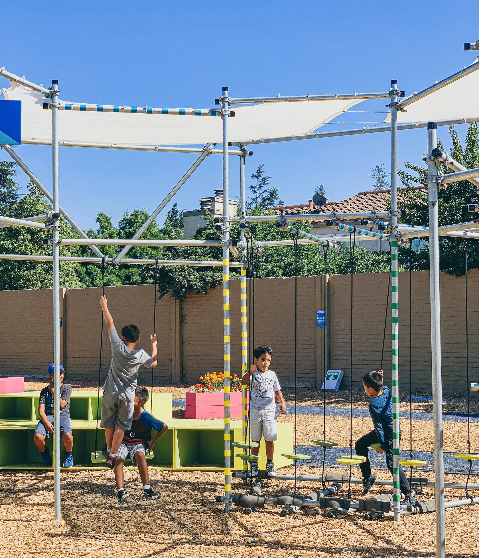 Three children try their luck on a ropes course, shaded from the summer sun by a scaffolding structure.