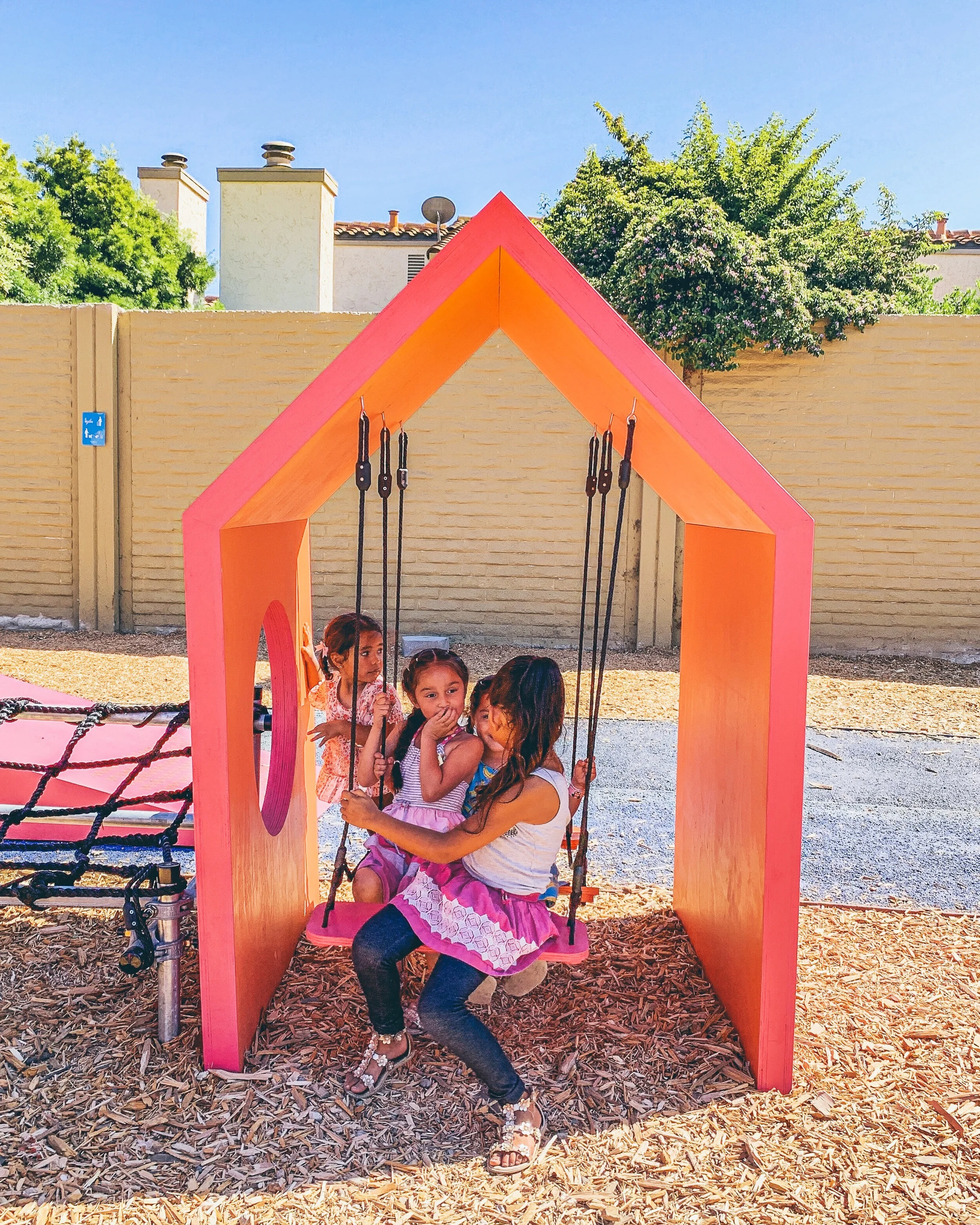 A simple house-like shape houses a set of swings, where three girls are in deep conversation.