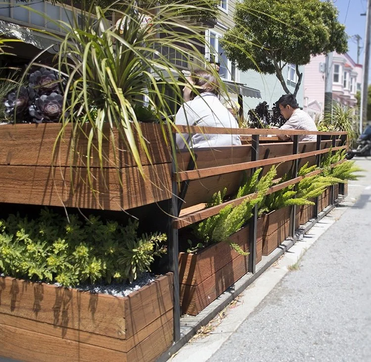 People enjoy their food at a parklet, complete with wooden planters, overflown with ferns and other plants.