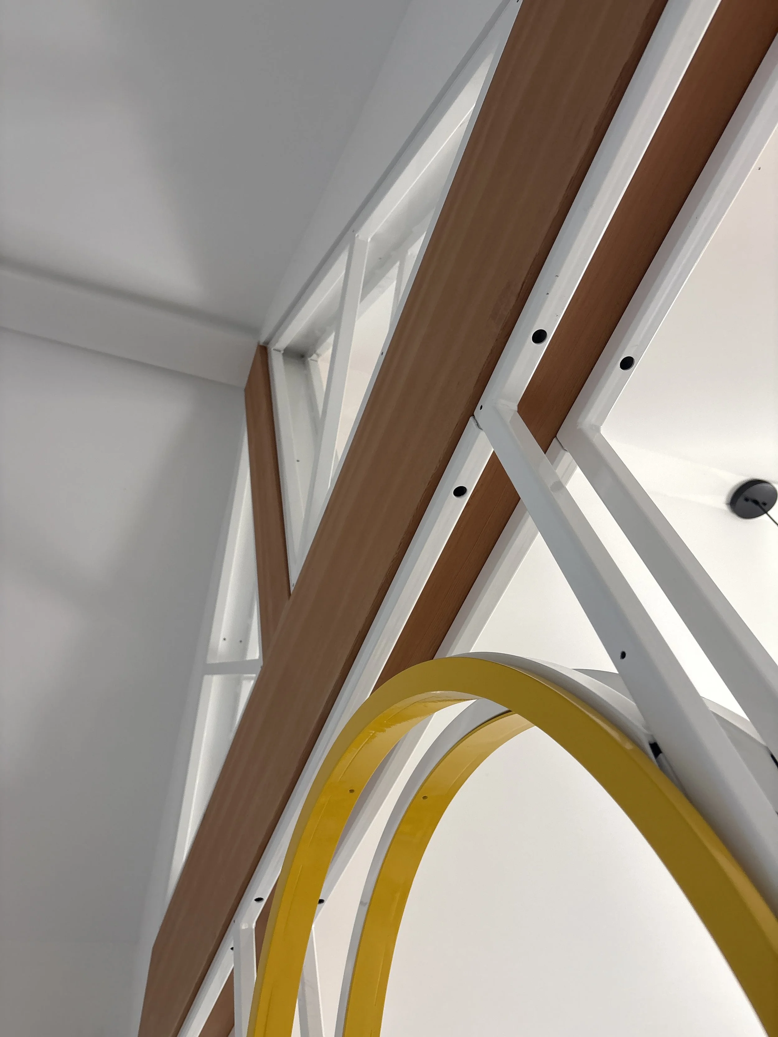 Close-up of a decorative wall next to a white shelf with wooden paneling, yellow curved element in the foreground, and a white wall with a black plug in the background.