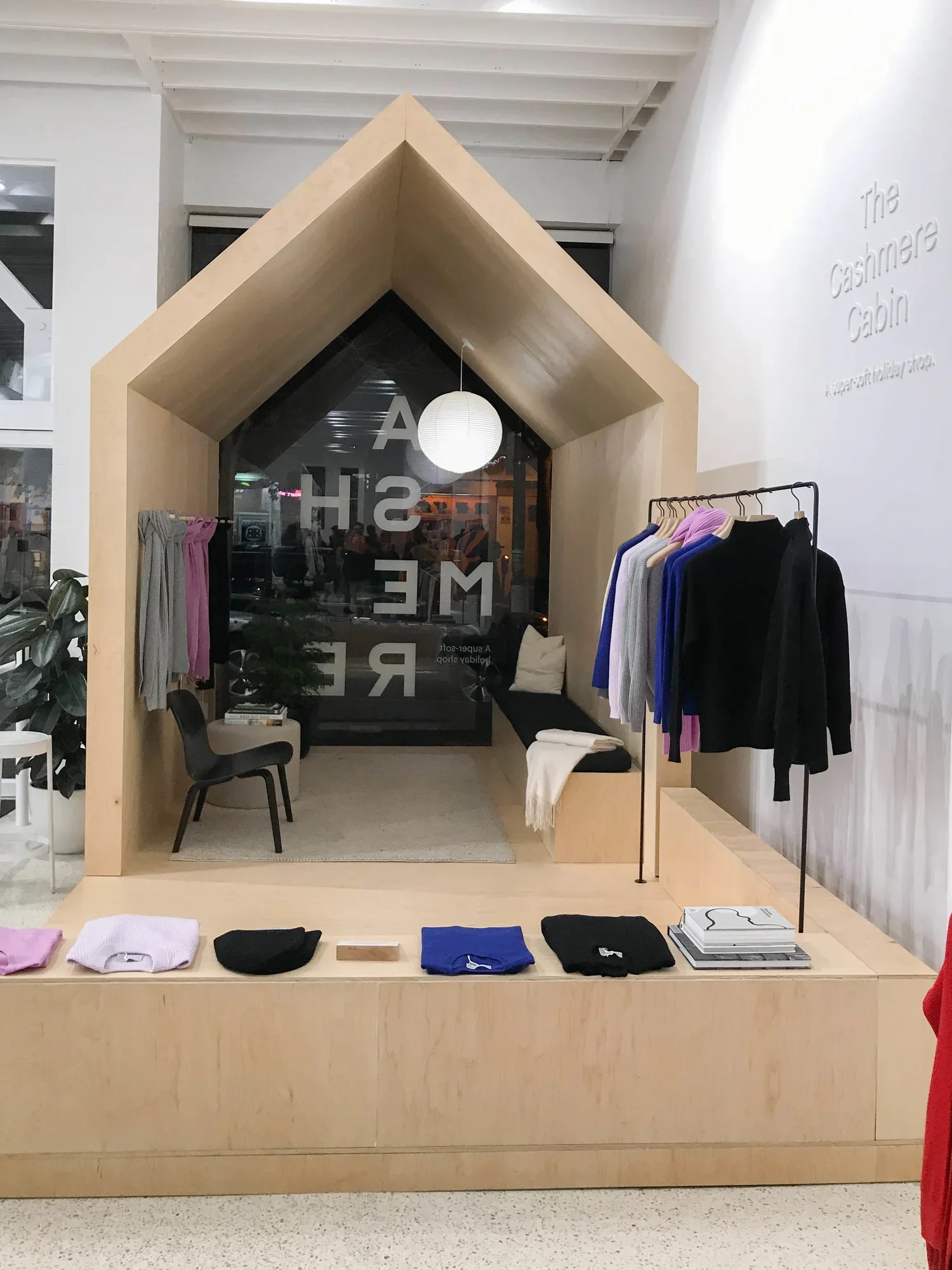 A retail display inside a clothing store features a wooden archway resembling a small house. Inside, there are clothes racks with sweaters and clothing items, and a selection of folded clothes on a table. The store has white walls and a large window with the store's name written on it. The right side of the image shows a rack with hanging hoodies or sweatshirts in various colors, including purple, gray, blue, and black.