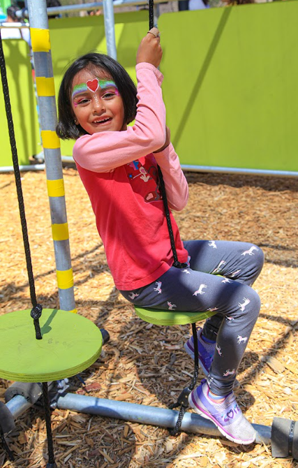 A young girl with face paint beams as she swings on a green swing at a playground.