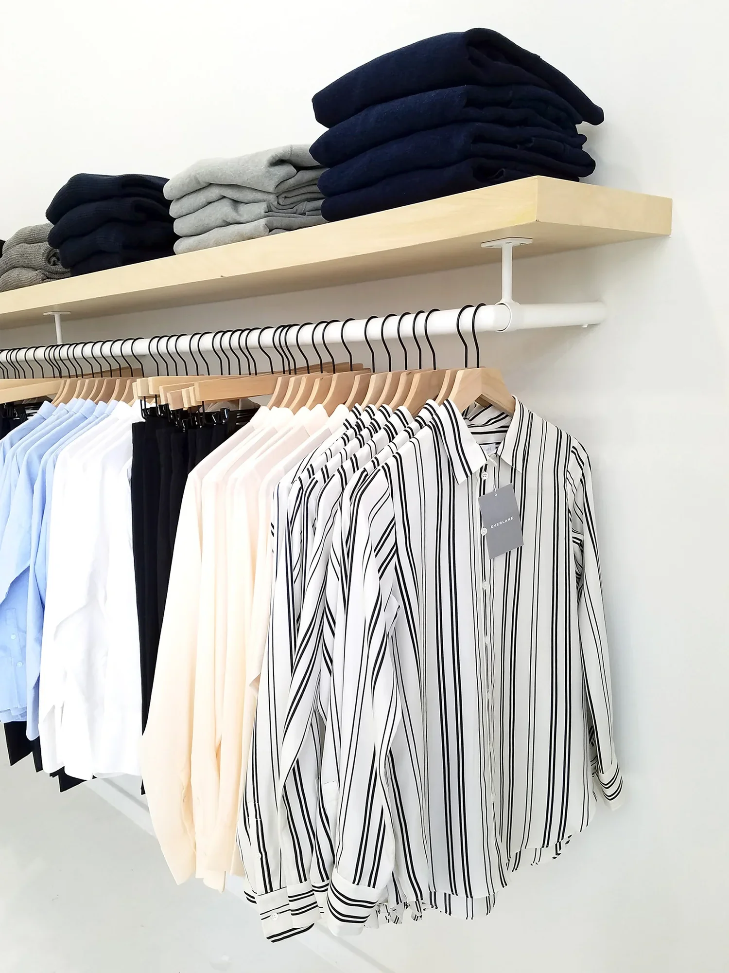Metal clothing rack with a wooden floating shelf above it hold mens shirts and sweaters.